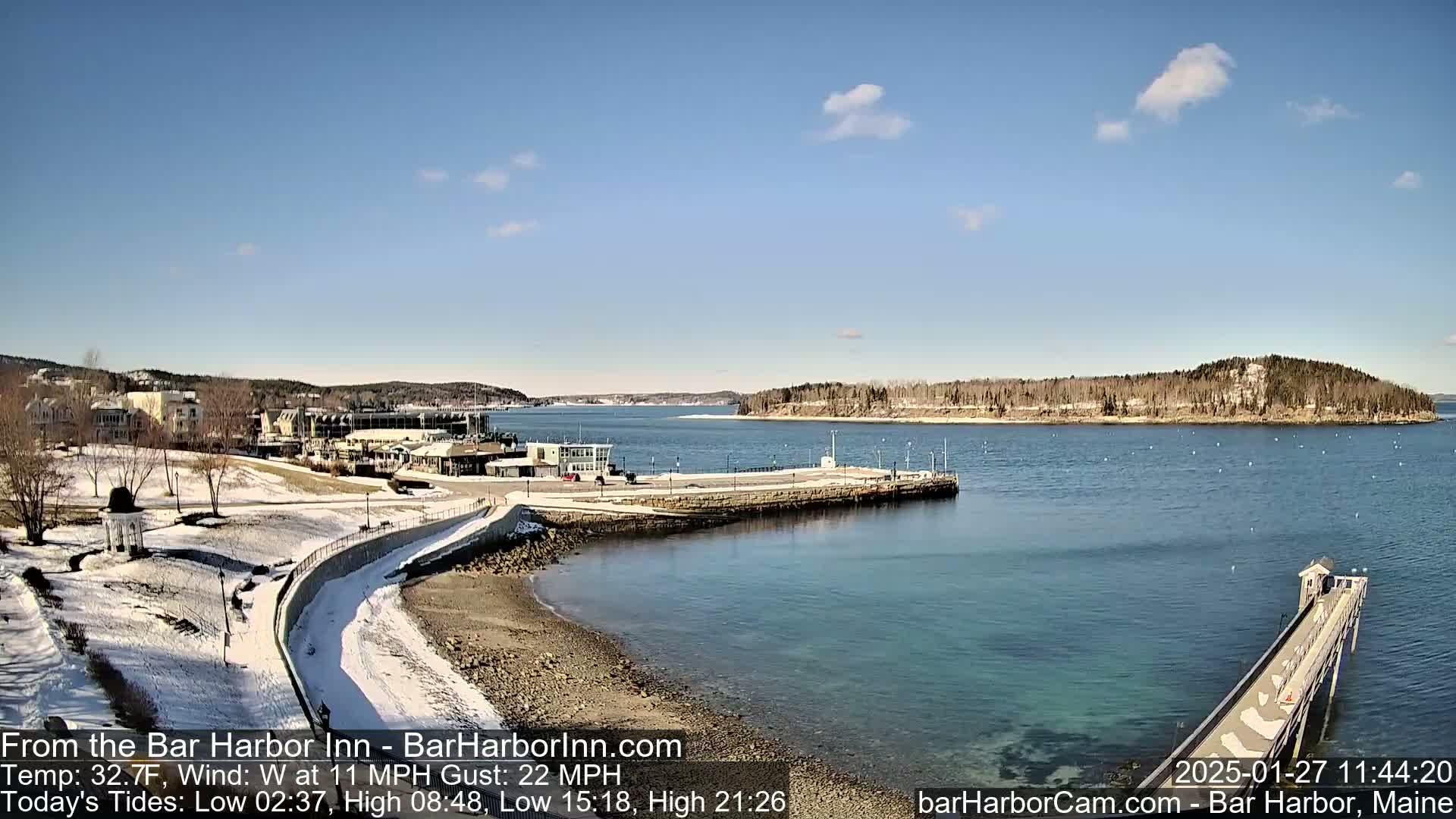 A partly sunny, cold day shows a snow-dusted waterfront scene with buildings, a long pier extending into a calm bay, and a small island in the distance.