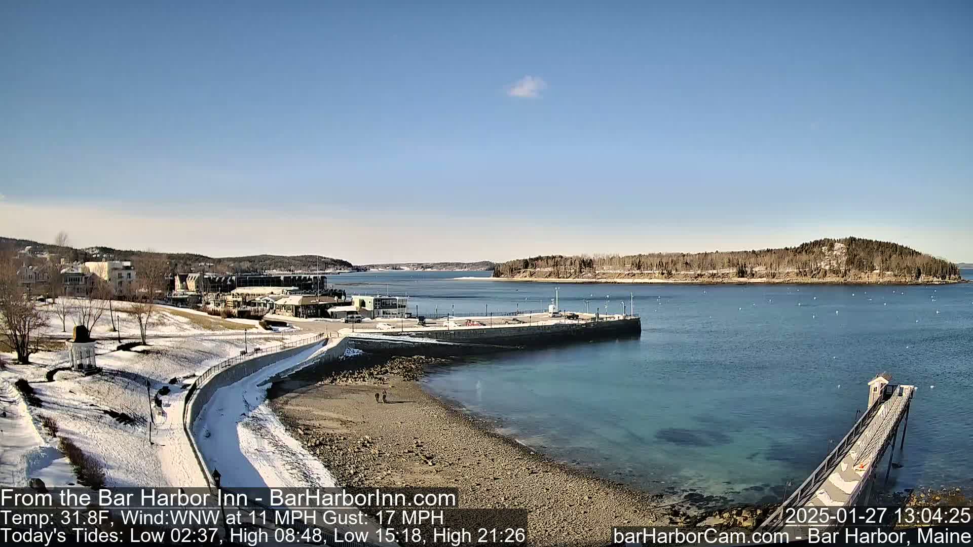 A snow-dusted coastal town with a harbor, a long pier, and a small island is visible under a clear blue sky.