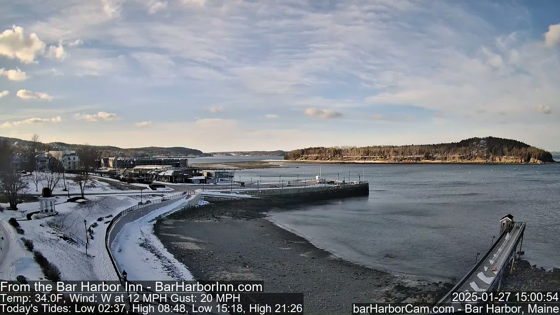 A snow-covered shoreline with a calm harbor, a pier, and buildings in the distance under a partly cloudy sky.
