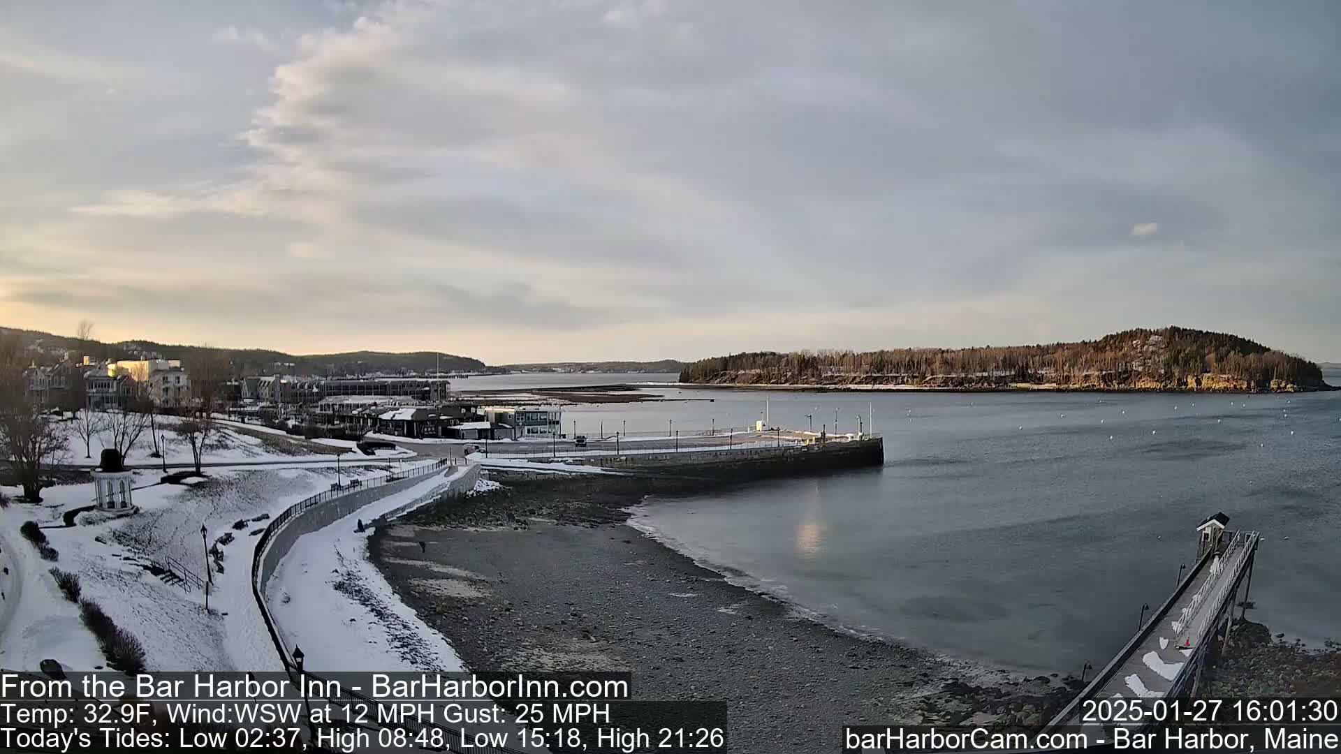 A snow-covered shoreline and buildings overlook a calm bay under a partly cloudy sky.