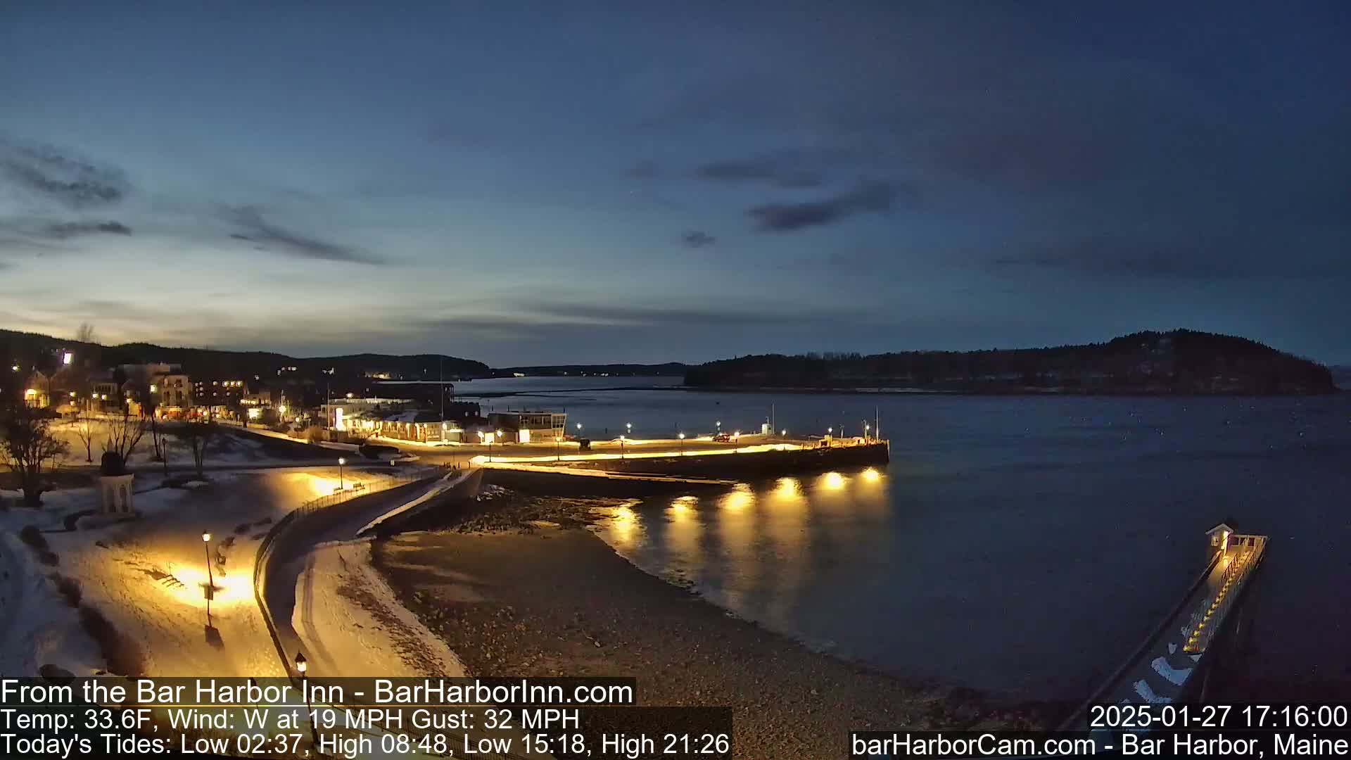 A snow-covered waterfront town at twilight, with a calm body of water reflecting the lights from a pier and buildings, under a mostly clear, dark blue sky.
