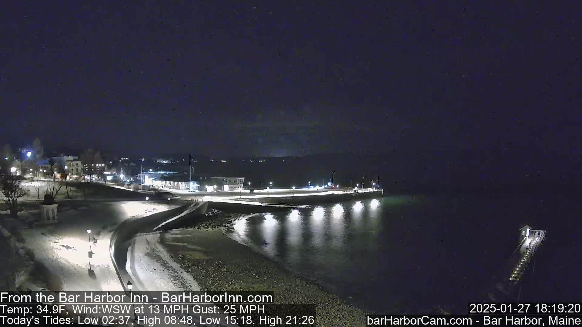 A snow-covered waterfront at night, illuminated by lights along a walkway and pier extending into calm, dark water.