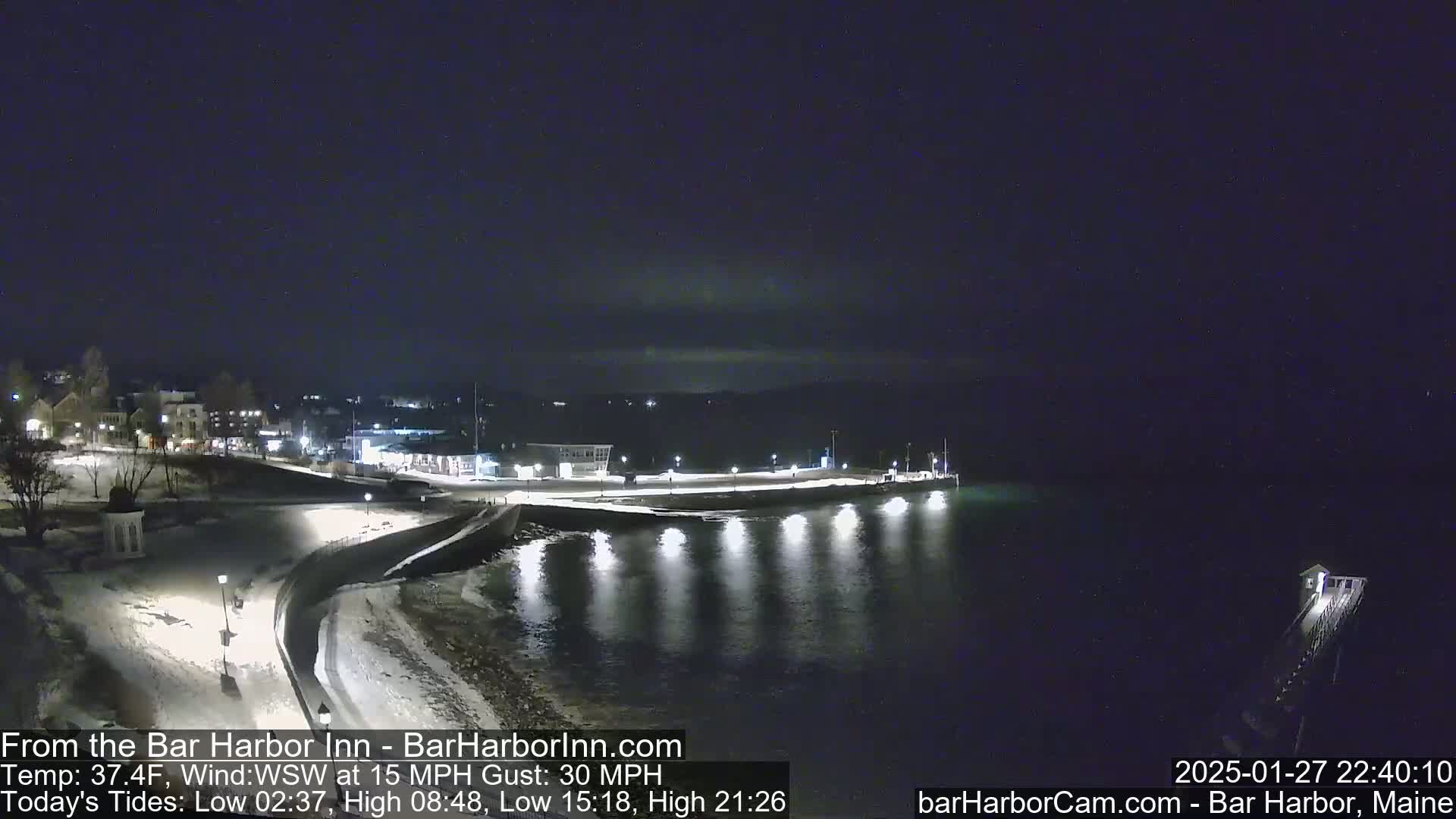 A nighttime view of a snow-covered waterfront with lights illuminating a calm body of water and a town in the background under a dark, clear sky.
