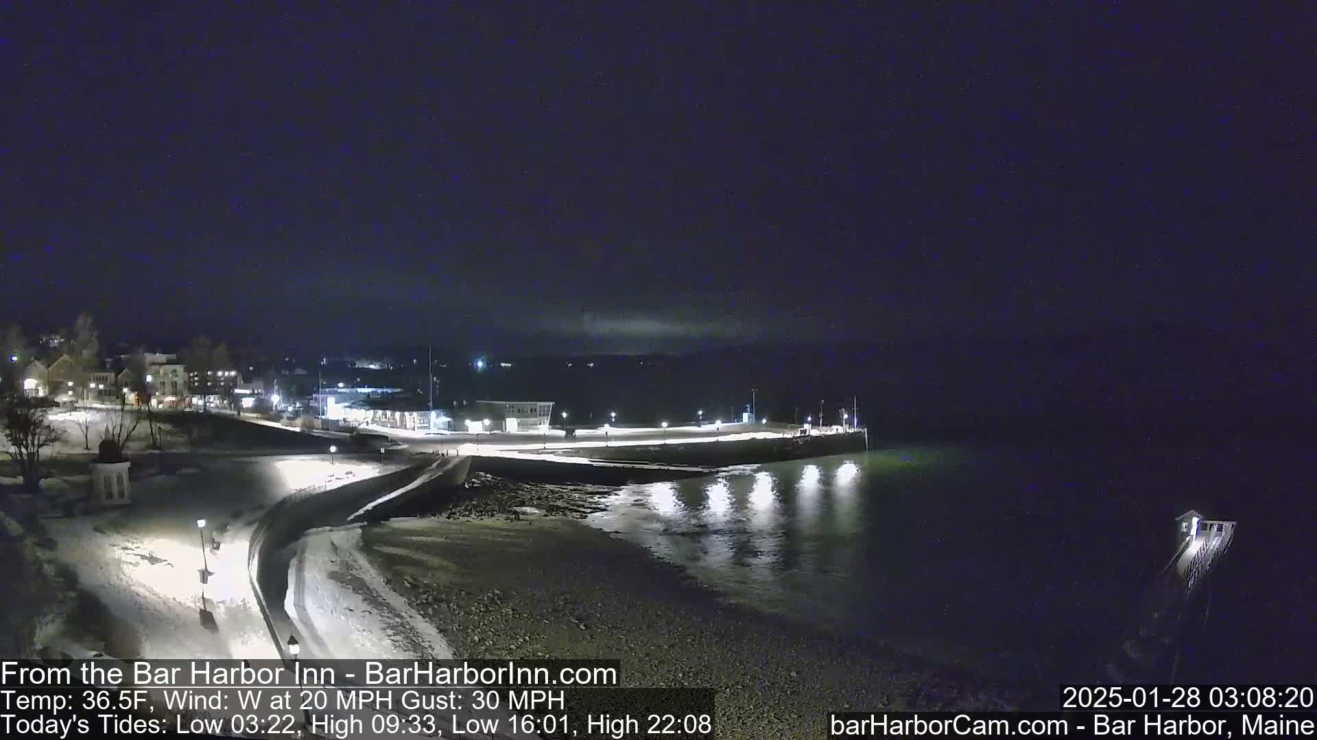 A nighttime view of a snow-covered shoreline with a town in the background, under a dark sky with a west wind at 20 mph and gusts up to 30 mph.