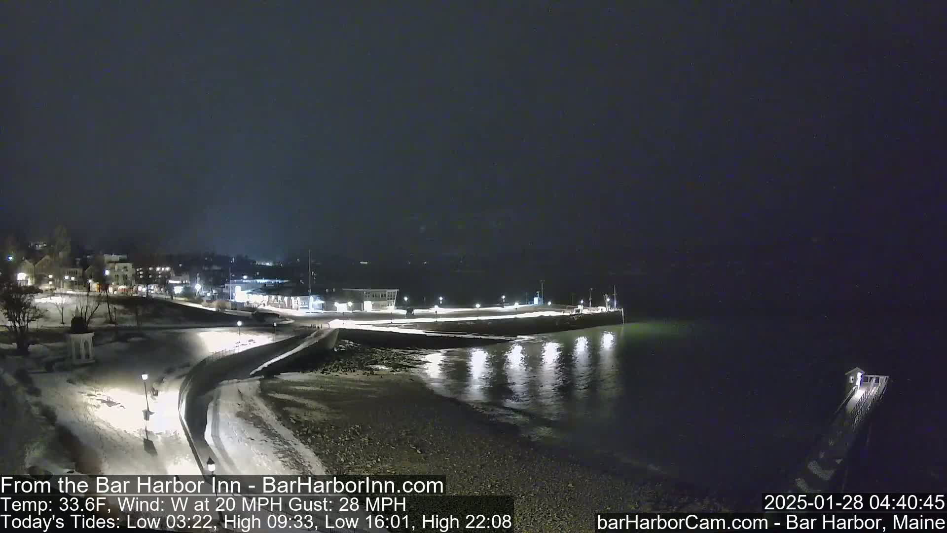 A snow-covered waterfront town is illuminated at night under a dark sky, with a calm body of water reflecting lights from the shore and a 20 mph westerly wind.