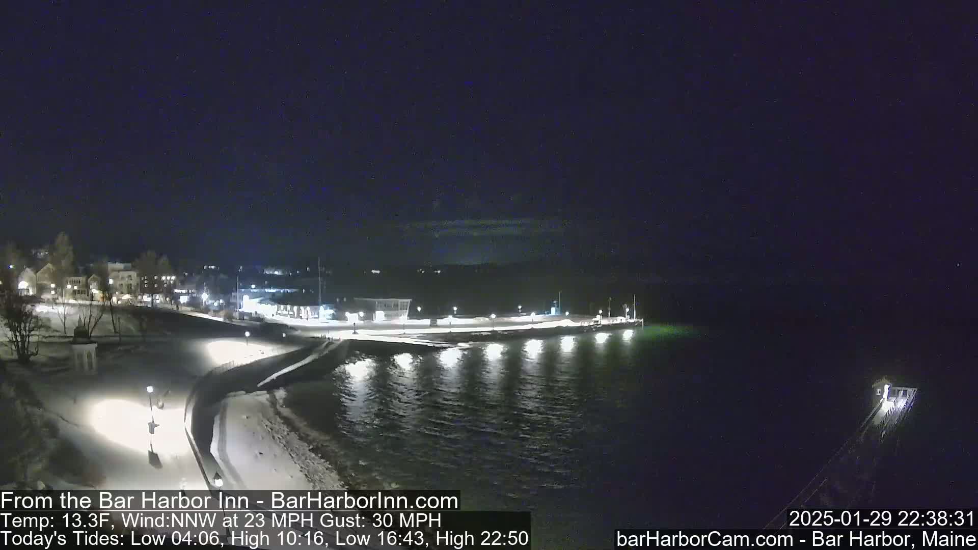 A snow-covered waterfront town at night, under a dark sky with a northwest wind of 23 mph and gusts up to 30 mph.