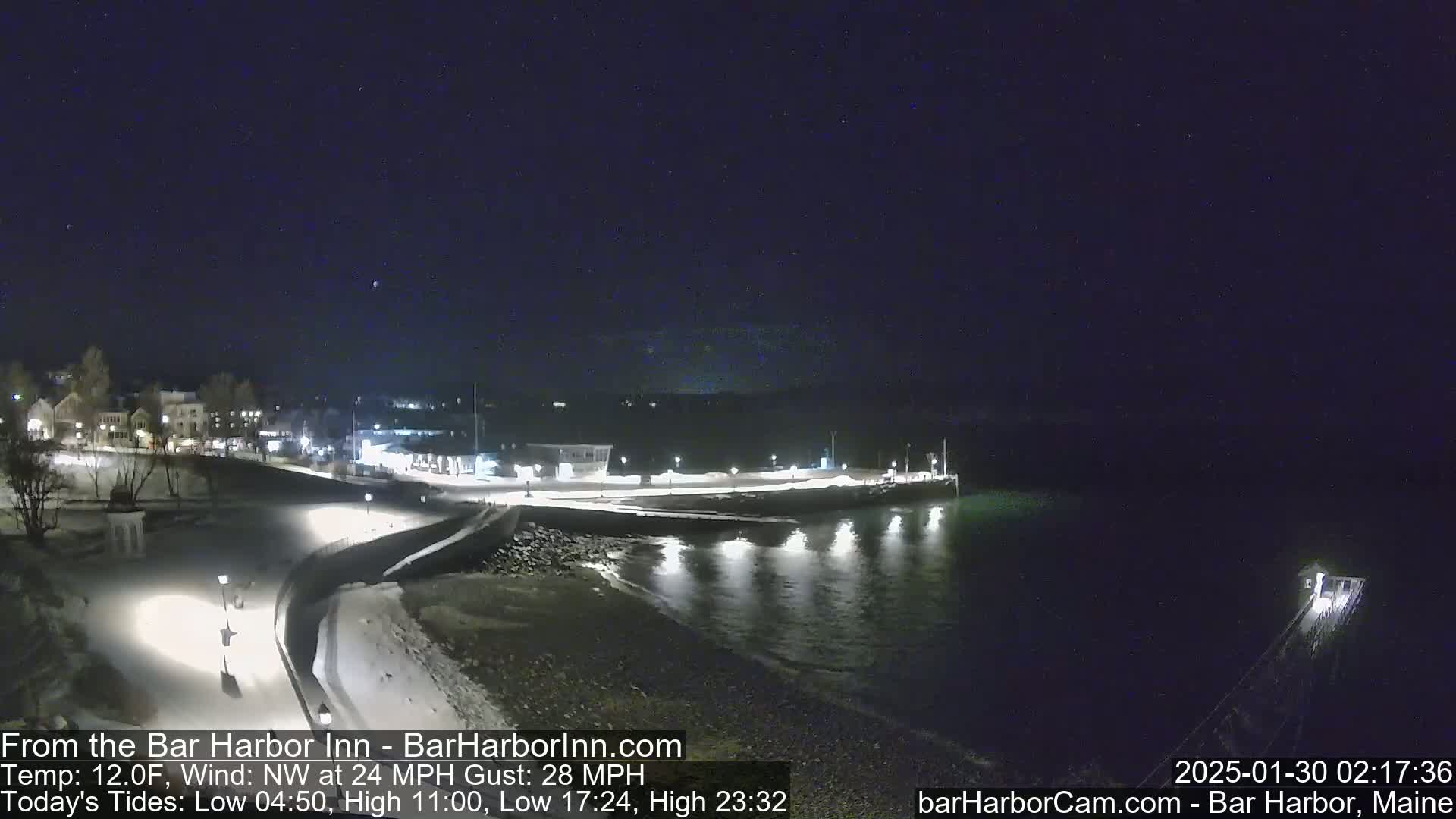 A snow-covered waterfront town at night, with calm water reflecting lights and a dark, clear sky.