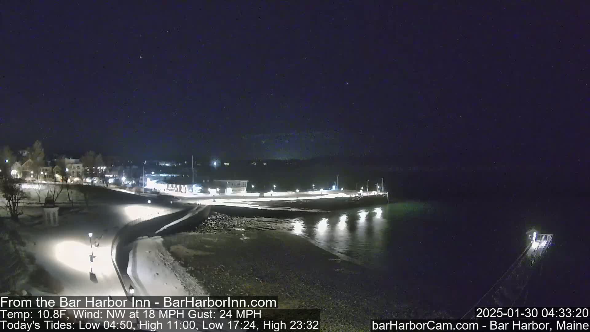 A nighttime view of a snow-covered waterfront town, illuminated by lights, with a dark, calm body of water under a clear, starry sky and a northwest wind.