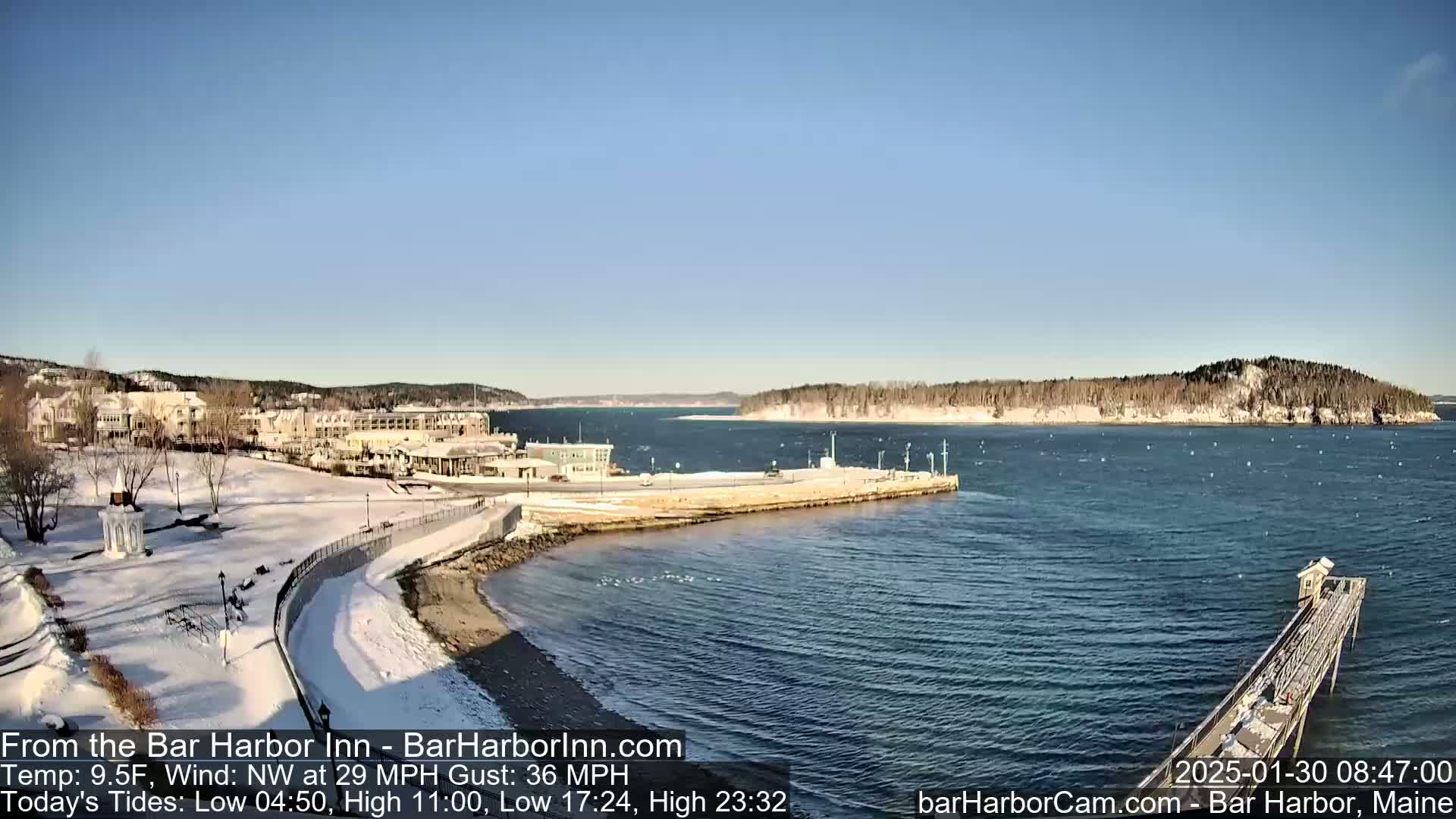 A snow-covered waterfront scene with a hotel and a long pier extending into a calm, blue ocean under a clear, sunny sky.