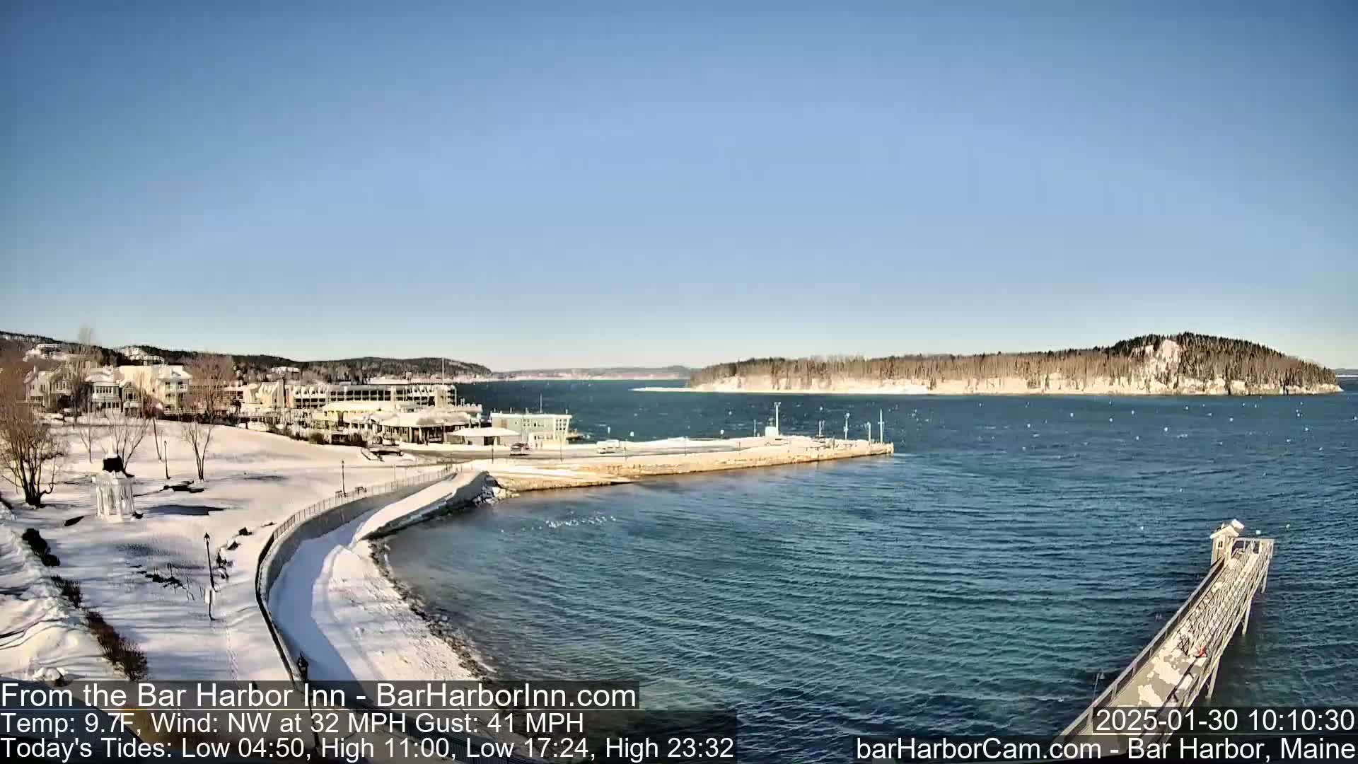 A snow-covered waterfront scene with a hotel and a long pier extending into a choppy ocean under a clear blue sky.
