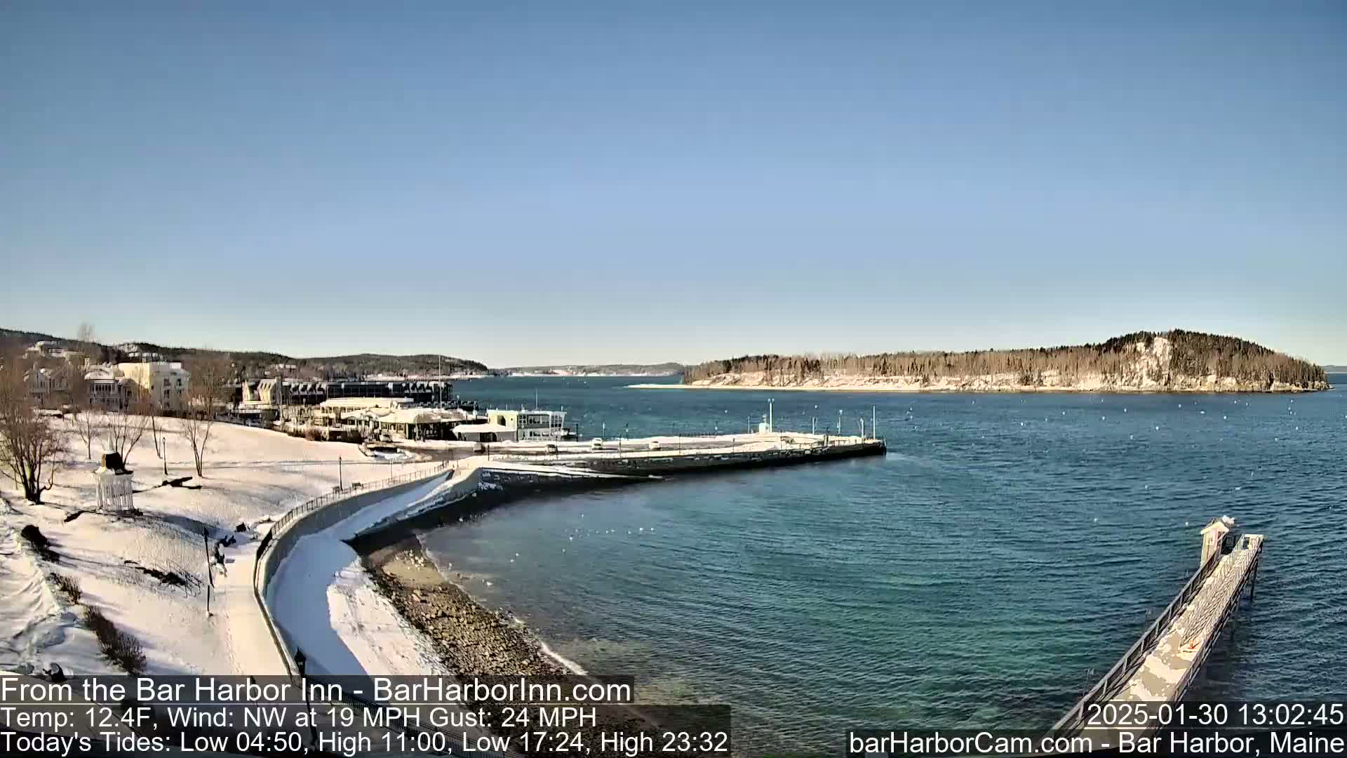 A snow-covered waterfront scene shows buildings, a partially frozen harbor, and a long pier extending into the water under a clear, sunny sky with a northwest wind.