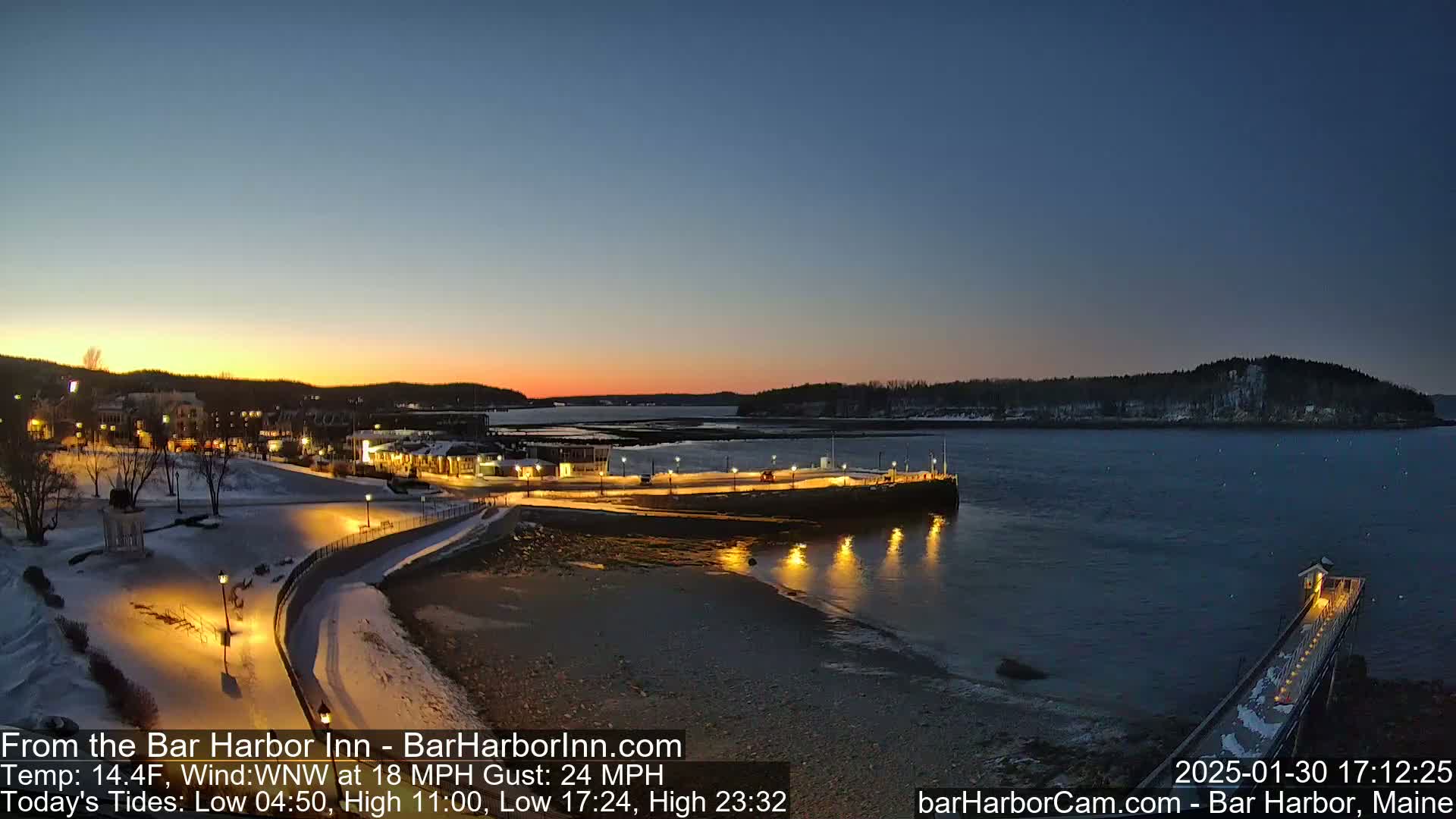 A snow-dusted waterfront town at twilight, with calm water reflecting lights from a pier and buildings, under a clear, dusky sky.