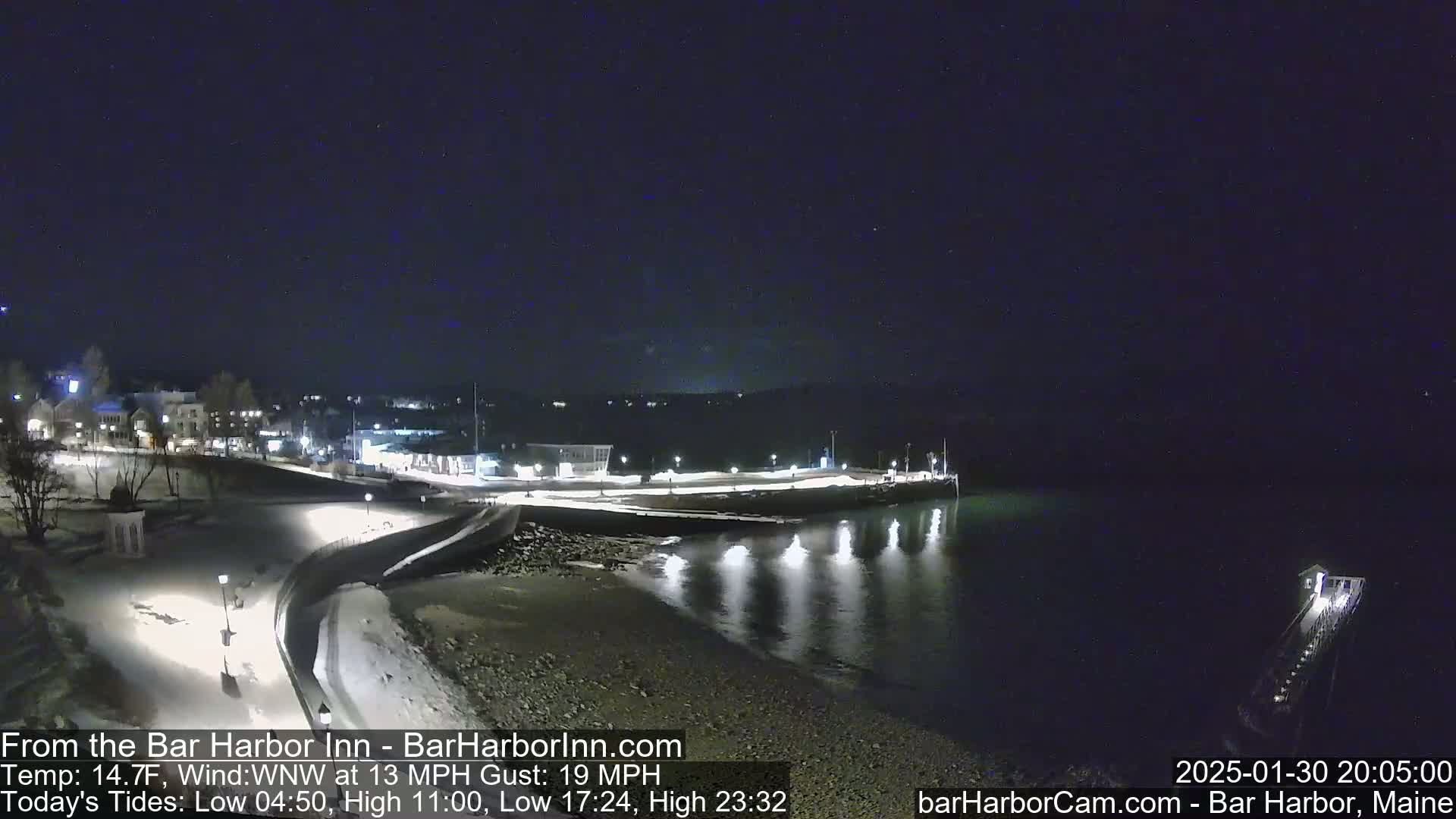 A snow-covered waterfront at night, with calm water reflecting lights from a nearby town under a clear, dark sky.