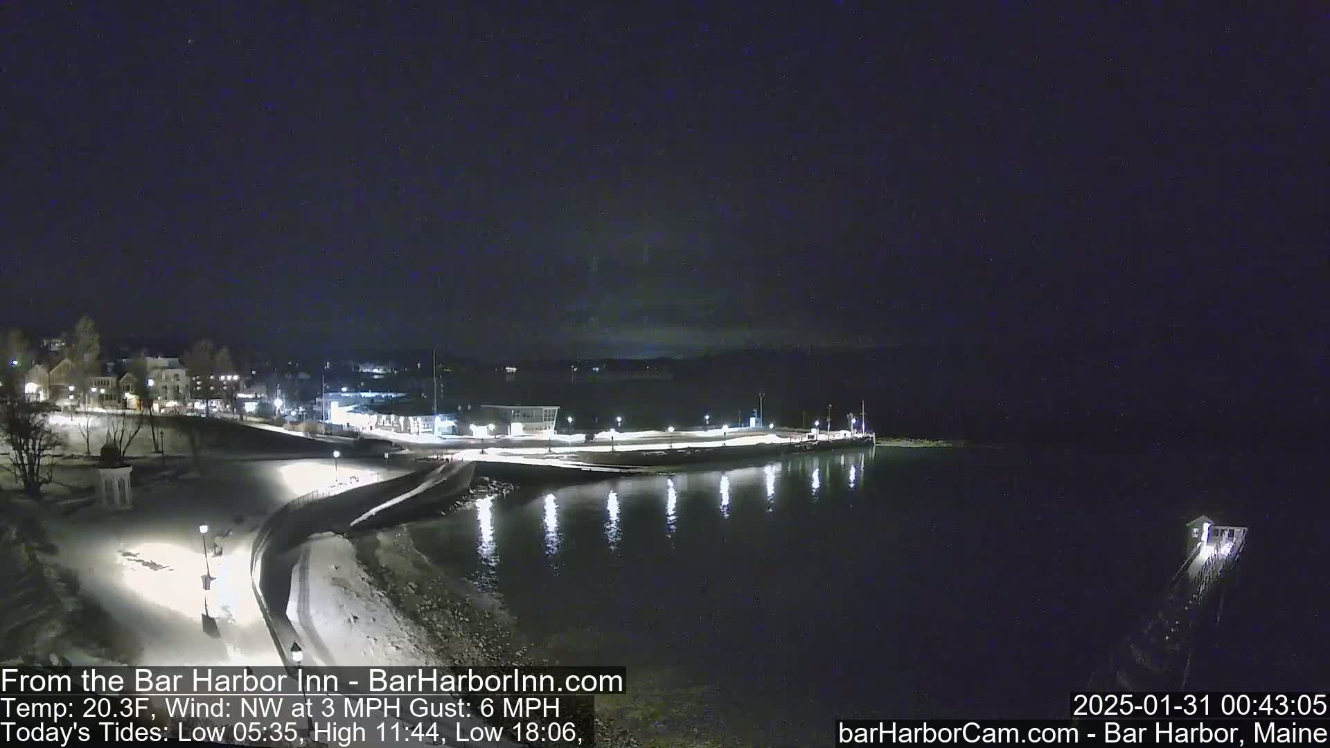 A nighttime, snow-covered waterfront scene shows a town's lights reflecting on calm water under a clear, dark sky.