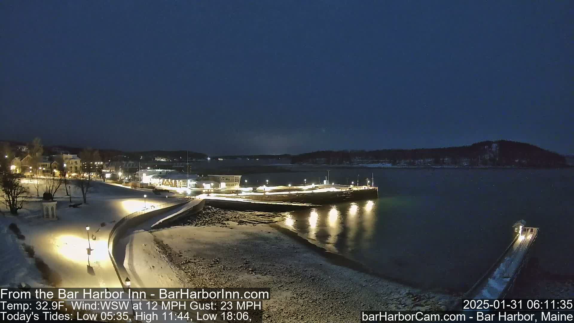 A snow-covered waterfront town is illuminated at night under a dark sky, with a calm body of water reflecting the lights.