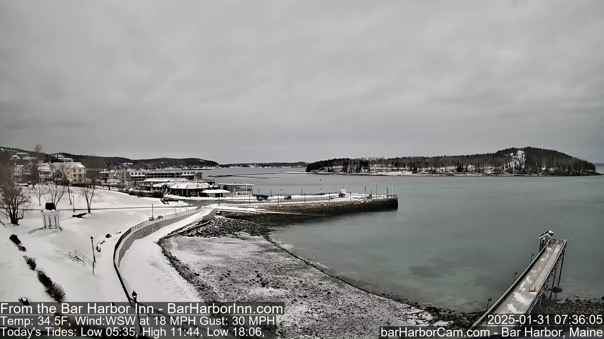 A snow-covered waterfront scene shows a calm harbor with a pier and a small town in the background under an overcast sky.