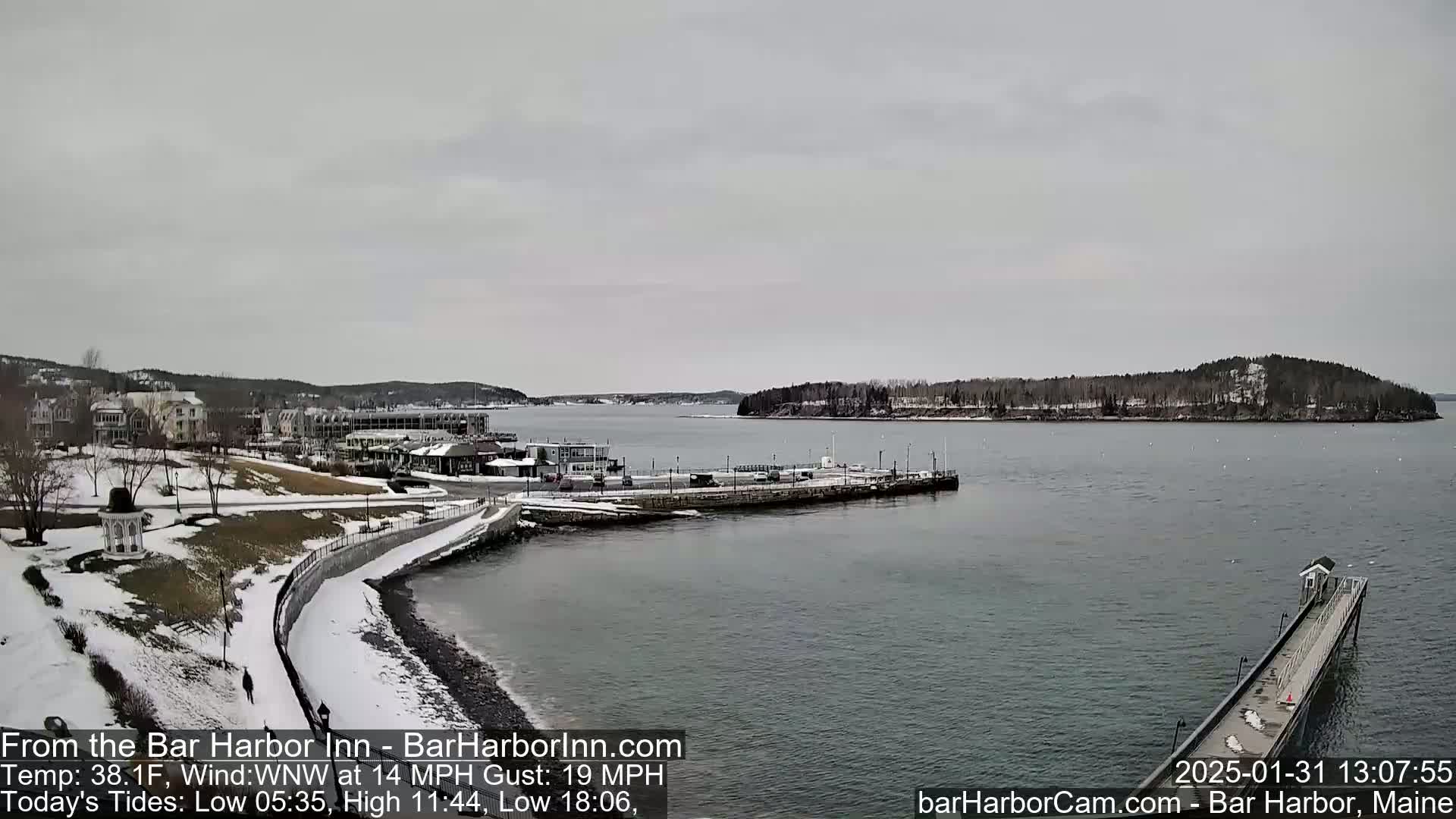 A snow-dusted coastal town overlooks a calm harbor with a long pier extending into the water under an overcast sky.