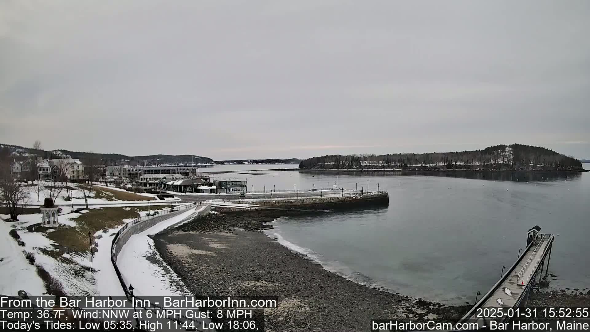 A snow-dusted coastal town and harbor are seen under a gray sky, with a partially icy shoreline and a long pier extending into the water.
