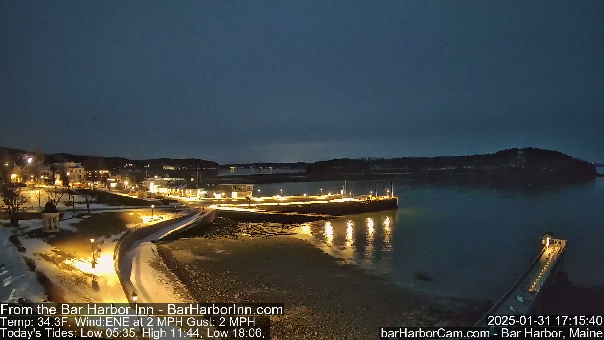 A nighttime view of a snow-covered harbor town with calm waters, lit walkways, and a pier extending into the dark water under a twilight sky.
