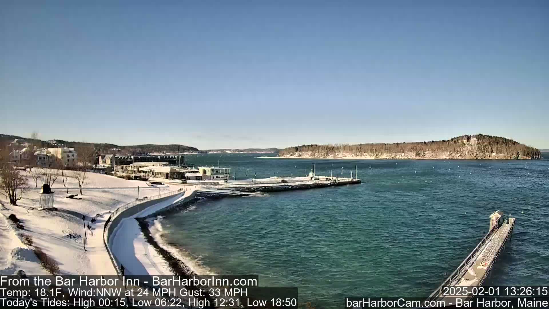 A snow-covered waterfront scene shows a partially frozen harbor with buildings and trees in the background under a clear, sunny sky.