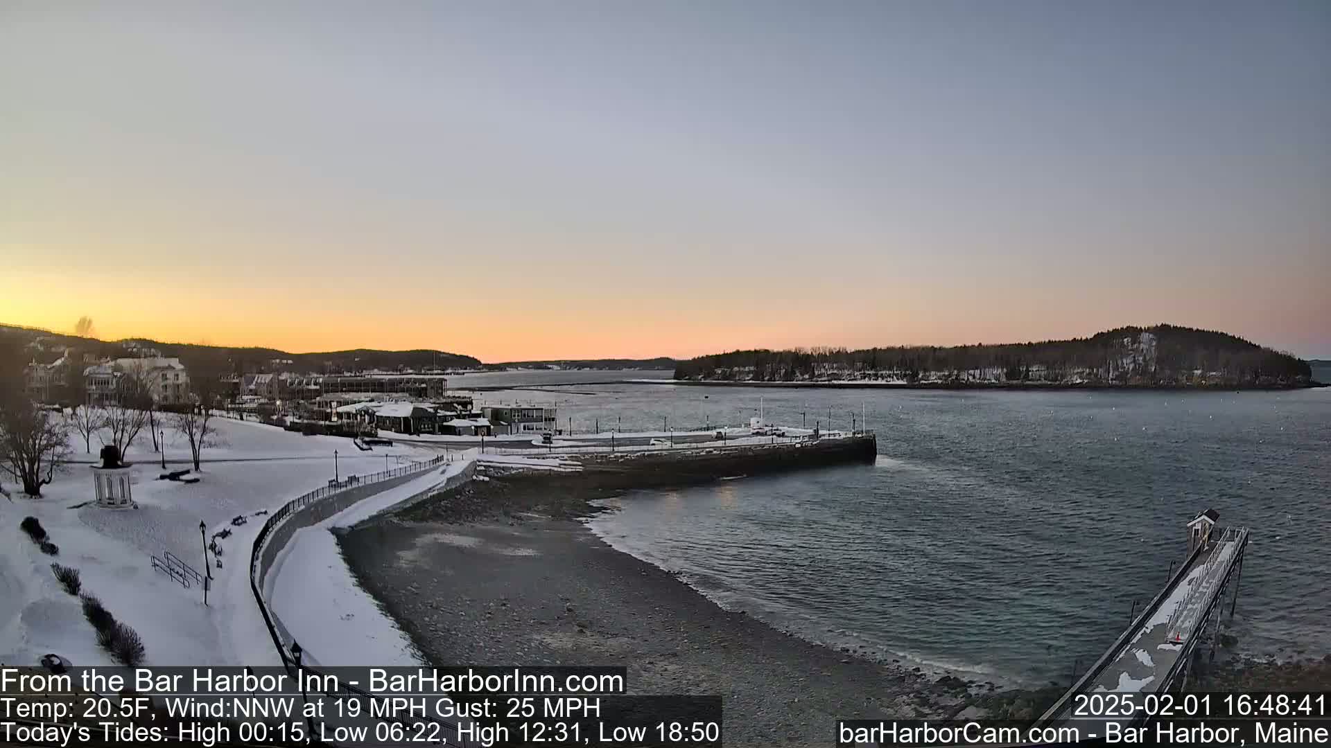 A snow-covered waterfront town at sunset, with calm water and a partly cloudy sky, shows a partially frozen harbor with a long pier extending into the water.