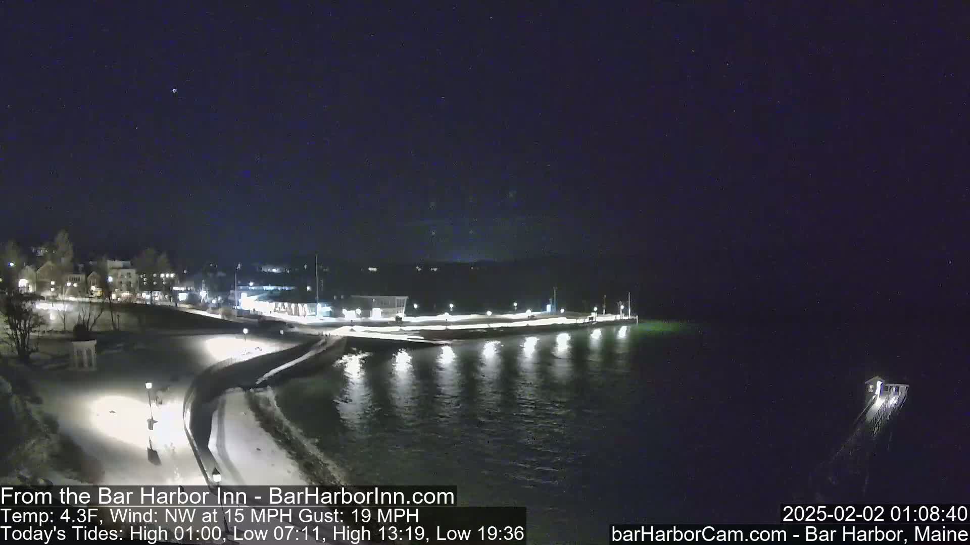 A nighttime view of a snow-covered waterfront, with calm water reflecting lights from buildings and a pier, under a clear, dark sky.