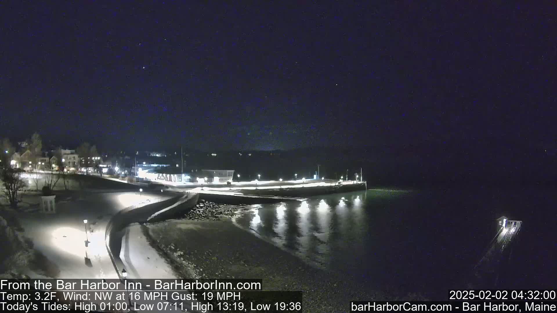 A snow-covered waterfront at night, under a clear, dark sky, with calm water reflecting lights from a nearby town.