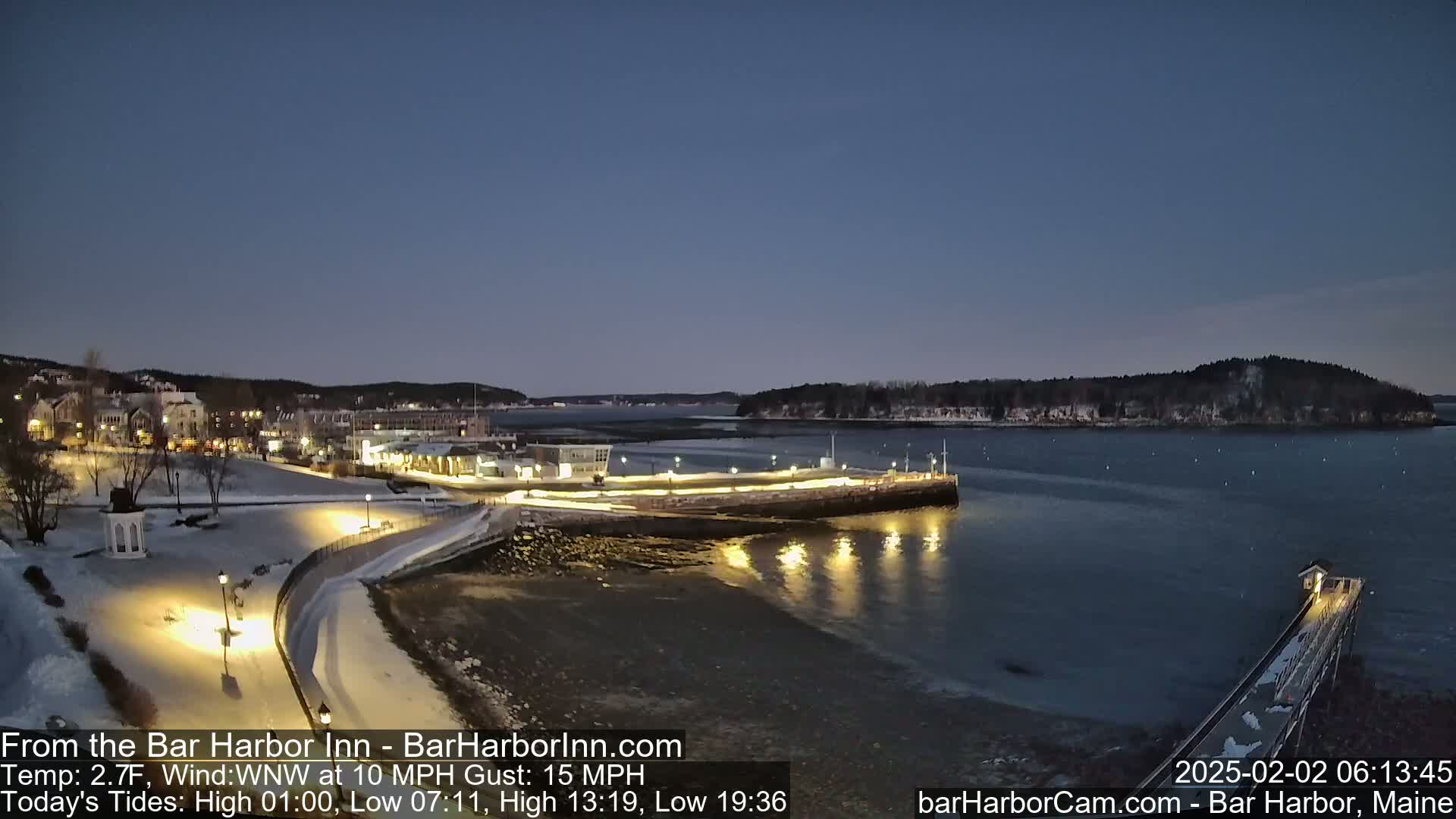 A snow-covered waterfront town at twilight, with calm water reflecting lights, under a clear, dark blue sky.