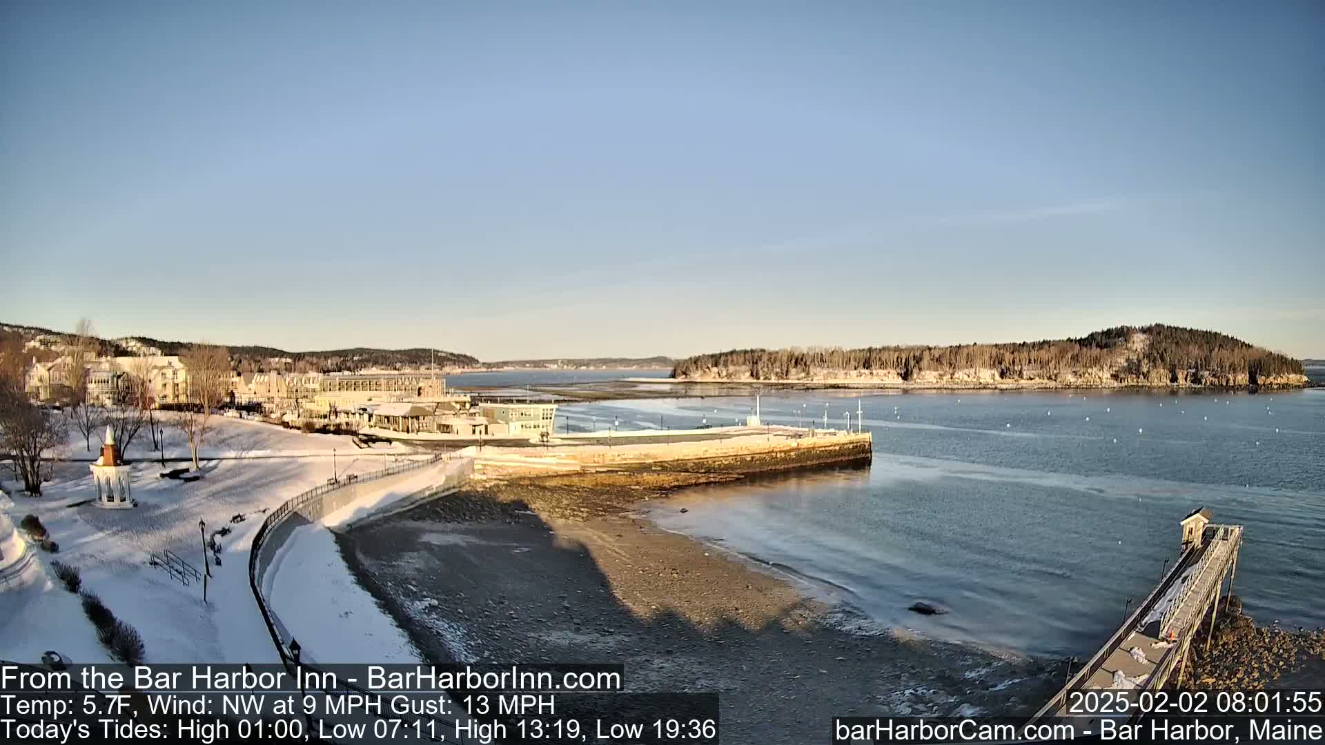 A snow-dusted coastal town with buildings and a partially frozen harbor is seen on a sunny, cold day.