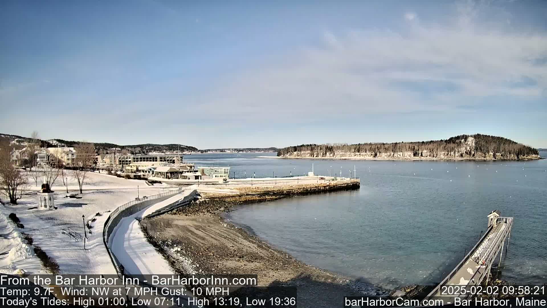 A snow-covered waterfront scene shows a calm bay with a long pier extending into the water, under a mostly clear sky.