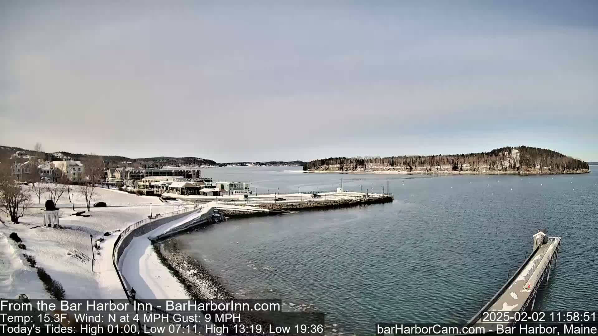 A snow-covered waterfront scene shows a harbor with buildings, a partially frozen shoreline, and a long pier extending into the calm water under a clear, bright sky.