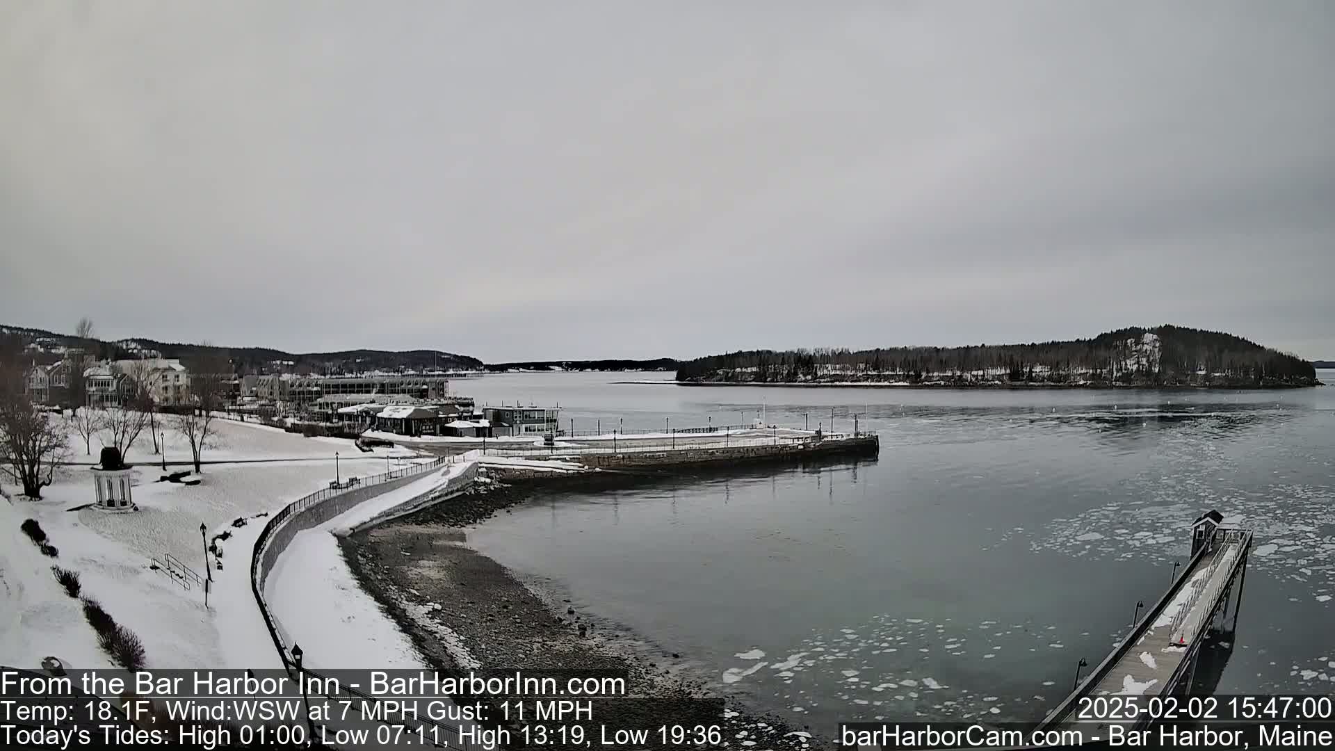 A snow-covered waterfront town with a calm harbor partially iced over under a cloudy sky.