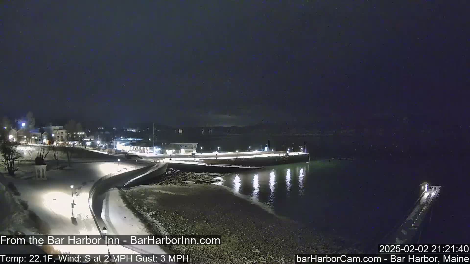 A snow-covered waterfront town at night, with calm water reflecting lights and a pier extending into the dark water under a clear, dark sky.