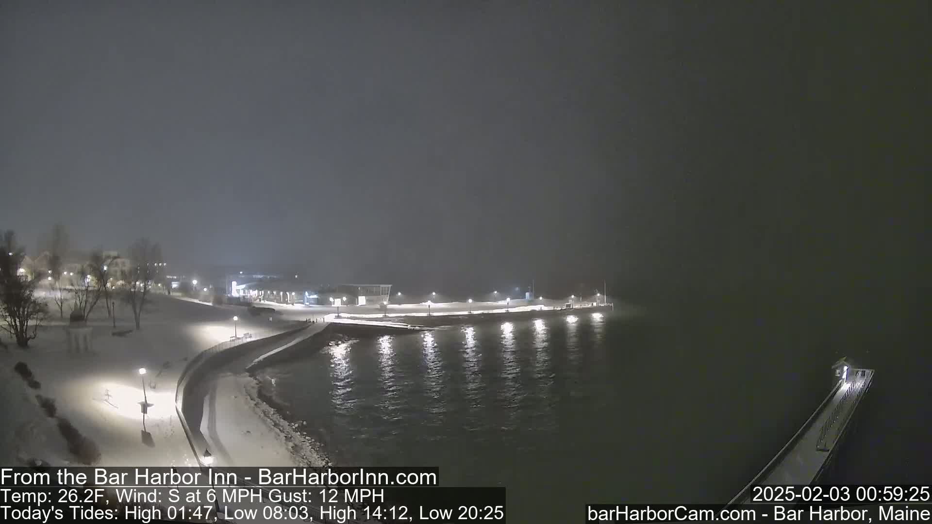A snow-covered waterfront at night with calm water, lights illuminating a snow-covered walkway and pier, under a hazy, dark sky.