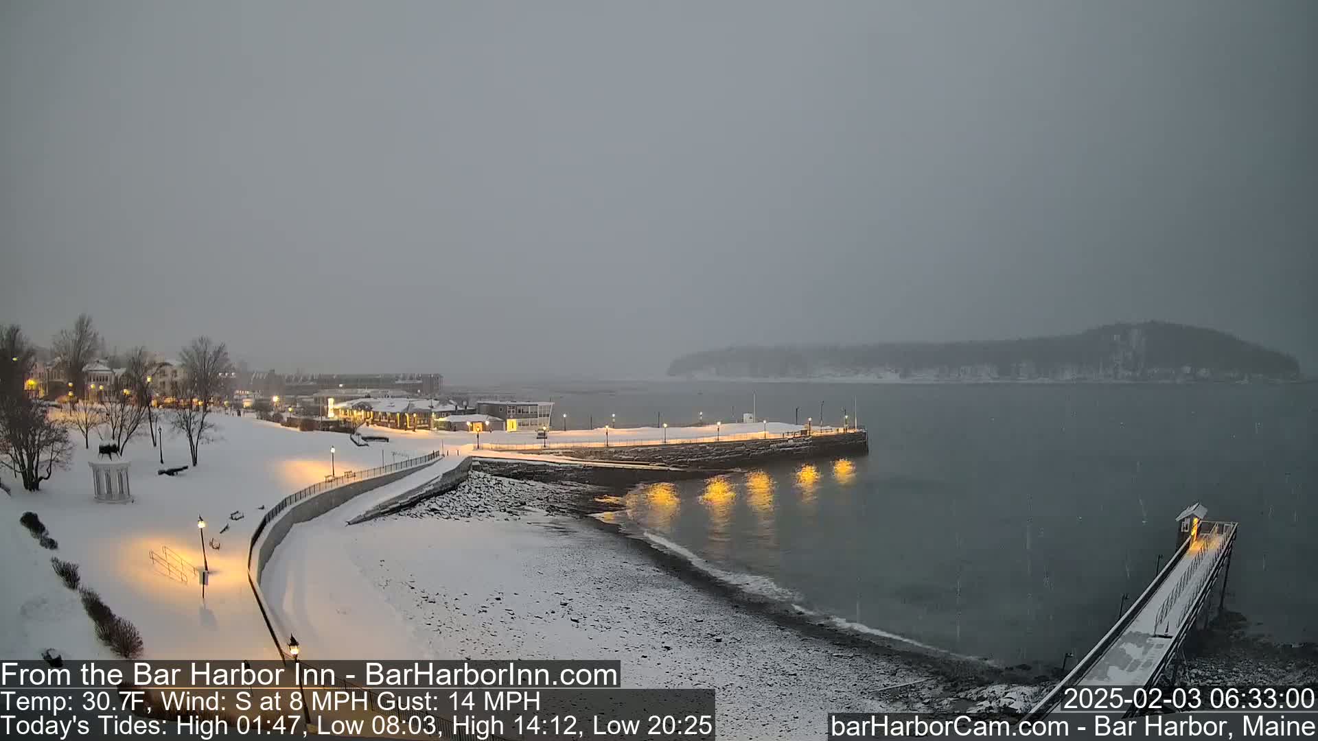 A snow-covered waterfront scene shows a town with lights on at night, a partially snow-covered pier extending into a dark body of water under a gray, overcast sky with light snowfall.