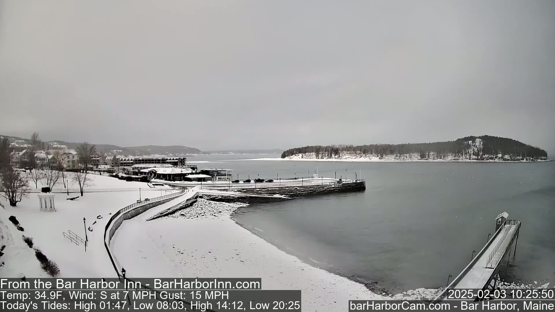 A snow-covered waterfront scene shows a partly frozen bay, a snow-covered town, and a pier extending into the calm water under an overcast sky.