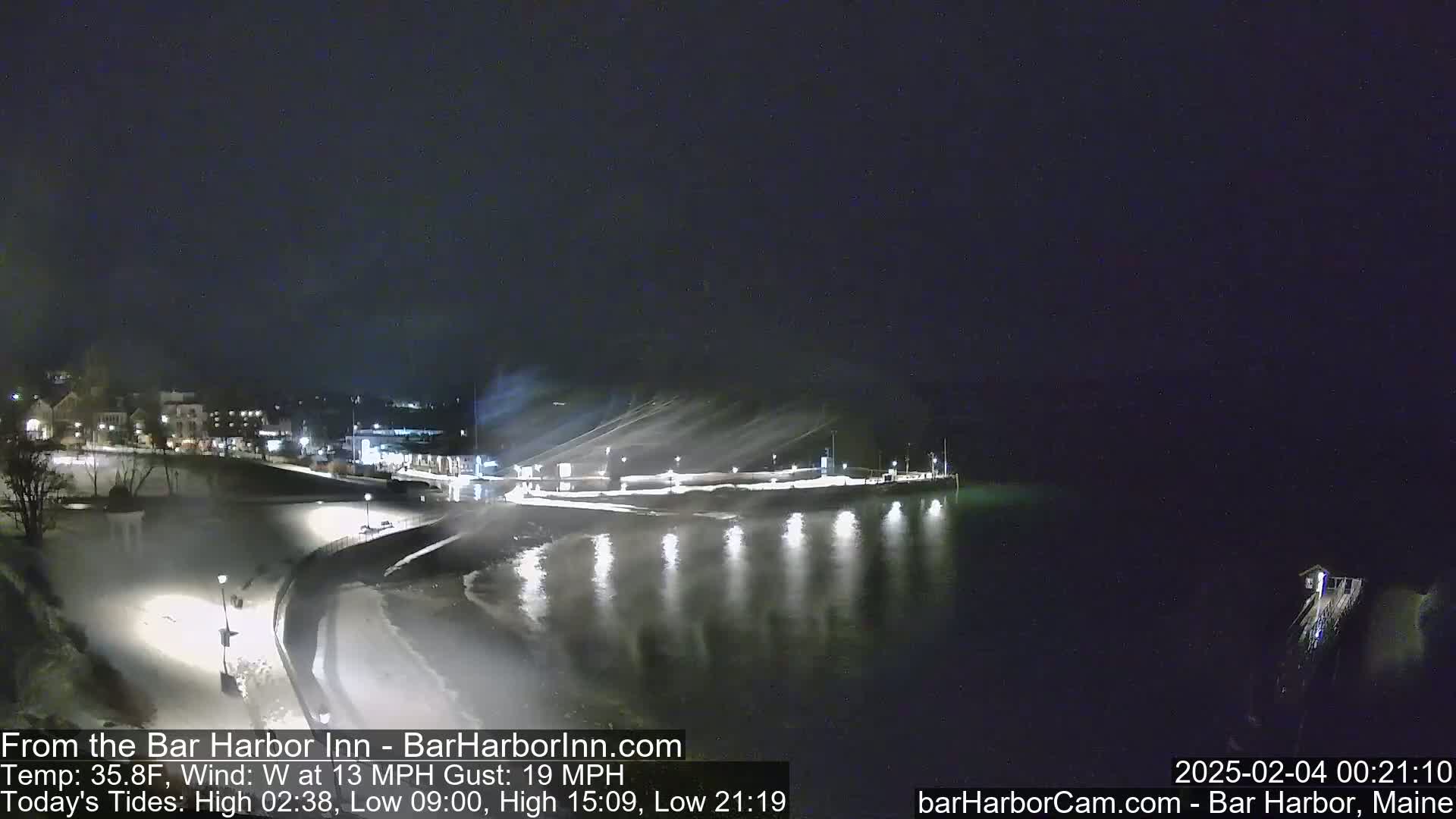 A nighttime, snowy scene shows a dark body of water reflecting lights from a snow-covered waterfront and buildings beyond.