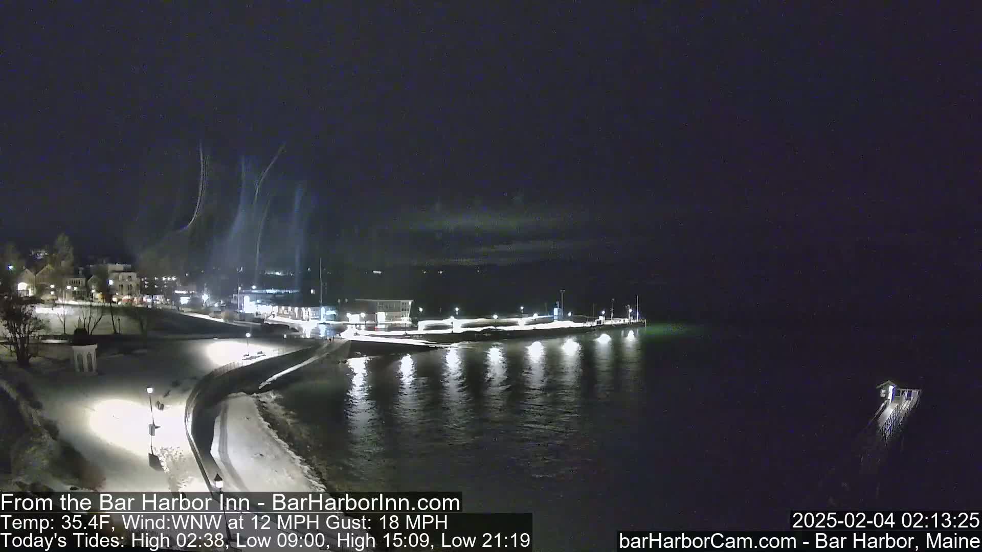 A nighttime, snow-covered waterfront scene shows a town lit up against a dark sky with faint streaks of light, possibly from snow blowing in the wind.