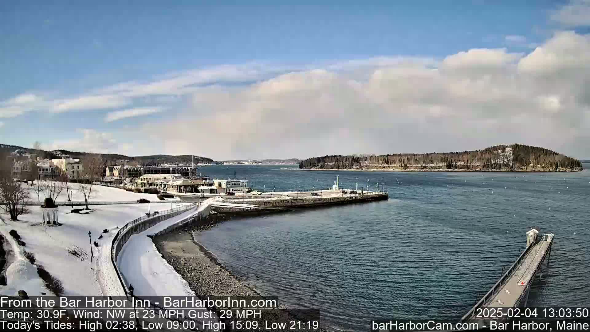 A snow-covered waterfront scene shows a harbor with buildings, a walkway, and a pier under a partly cloudy sky.