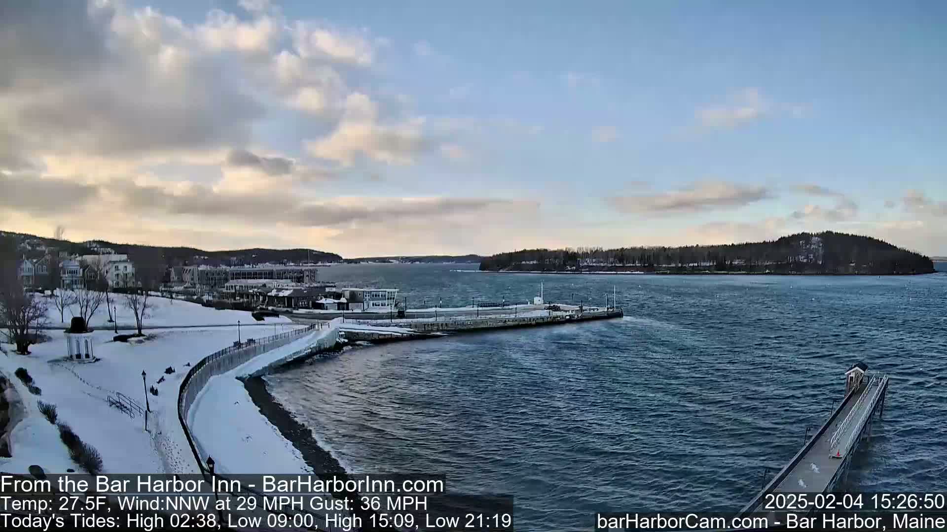 A snow-covered waterfront scene shows a calm body of water with a pier extending into it, buildings and trees along the shore under a partly cloudy sky.