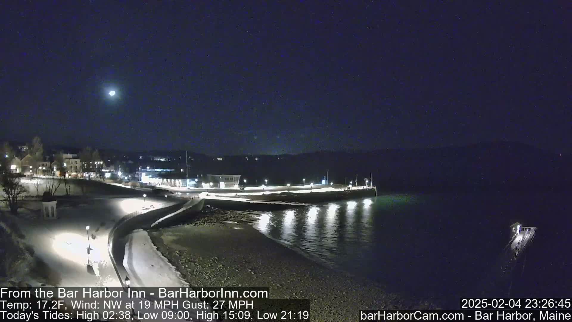 A nighttime coastal scene shows a snow-covered waterfront with buildings in the background under a clear sky and a bright moon, with a northwest wind at 19 mph and gusts up to 27 mph.