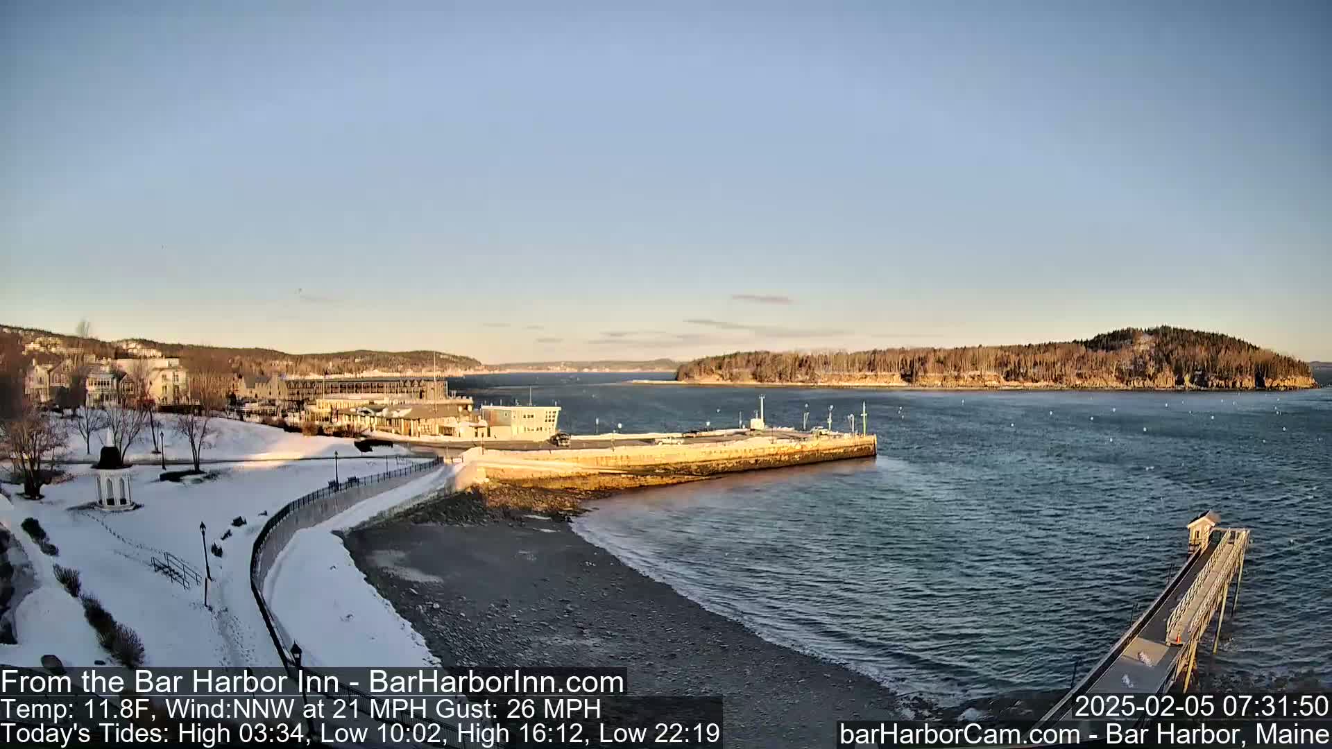 A snow-covered waterfront scene shows a harbor with buildings, a pier, and calm water under a clear, light blue sky.