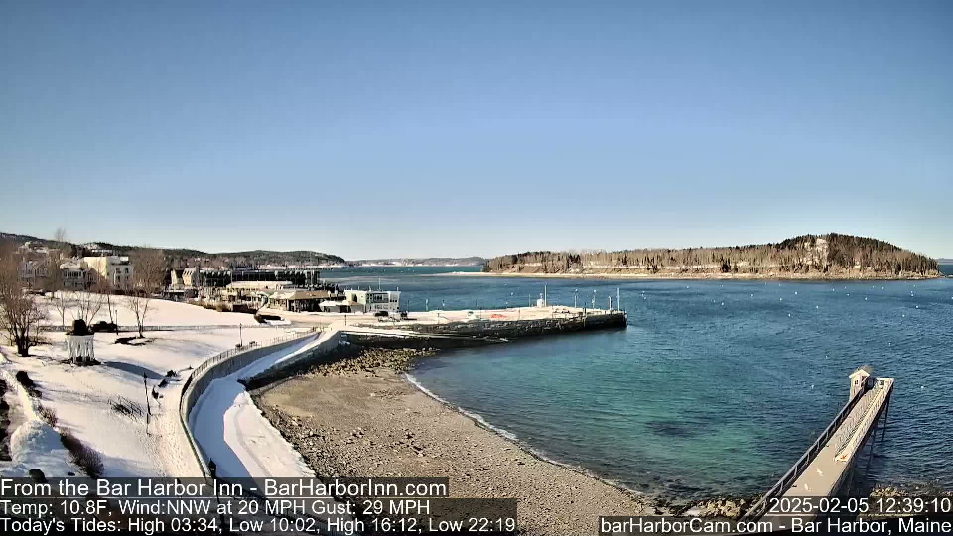 A snow-covered coastal town is seen on a sunny day with a calm harbor, and a long pier extending into the water.