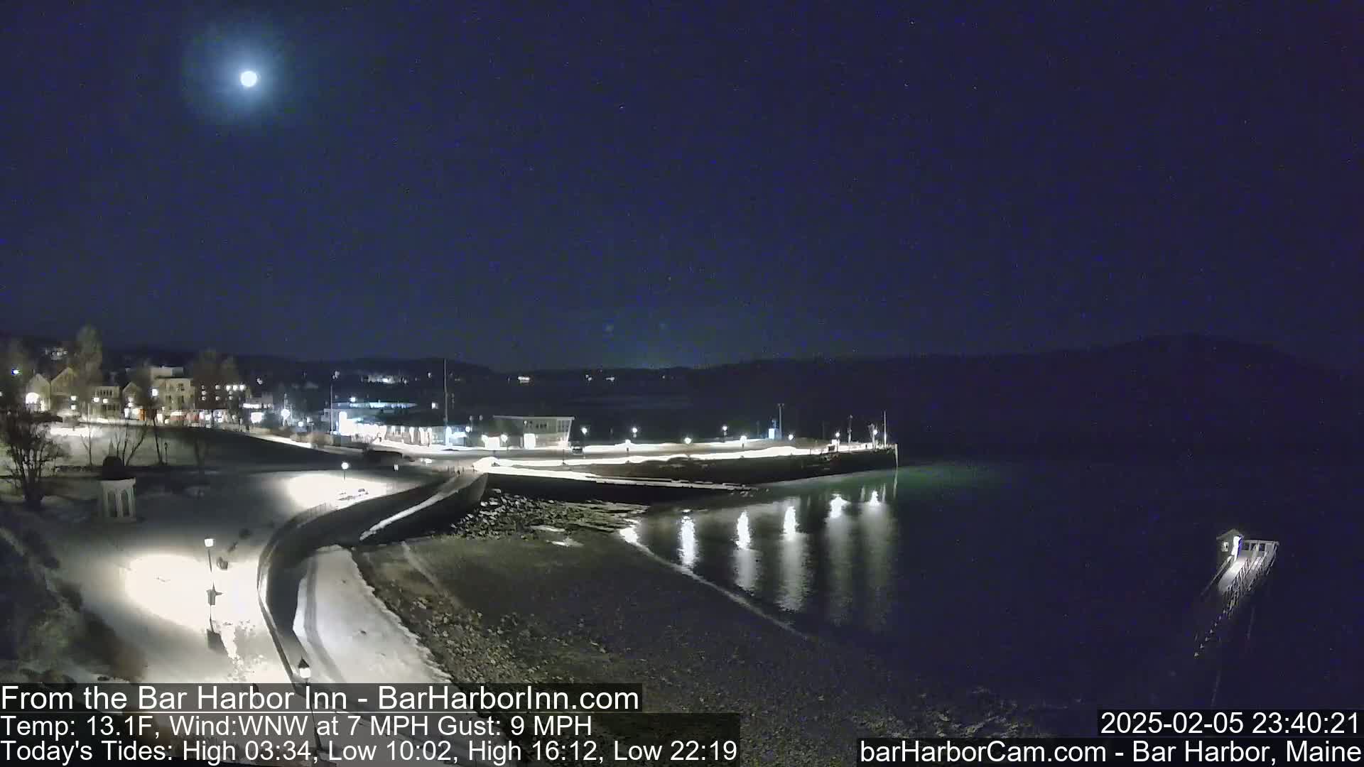 A nighttime view of a snow-covered harbor town under a bright moon, with calm water reflecting lights.