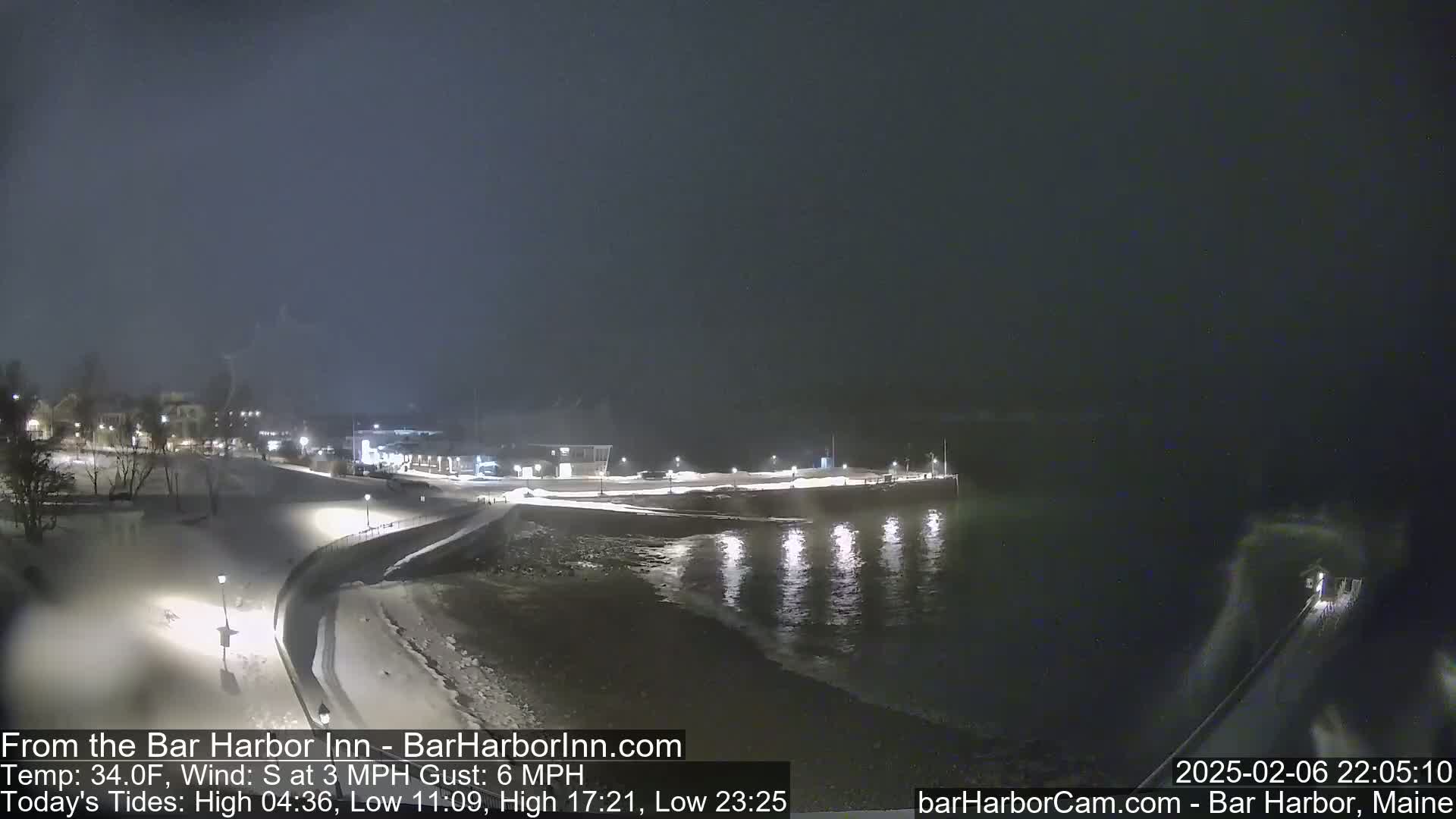 A nighttime snow-covered waterfront scene shows a dark body of water lapping at a snow-covered shore, with lights illuminating a pathway and buildings in the background.