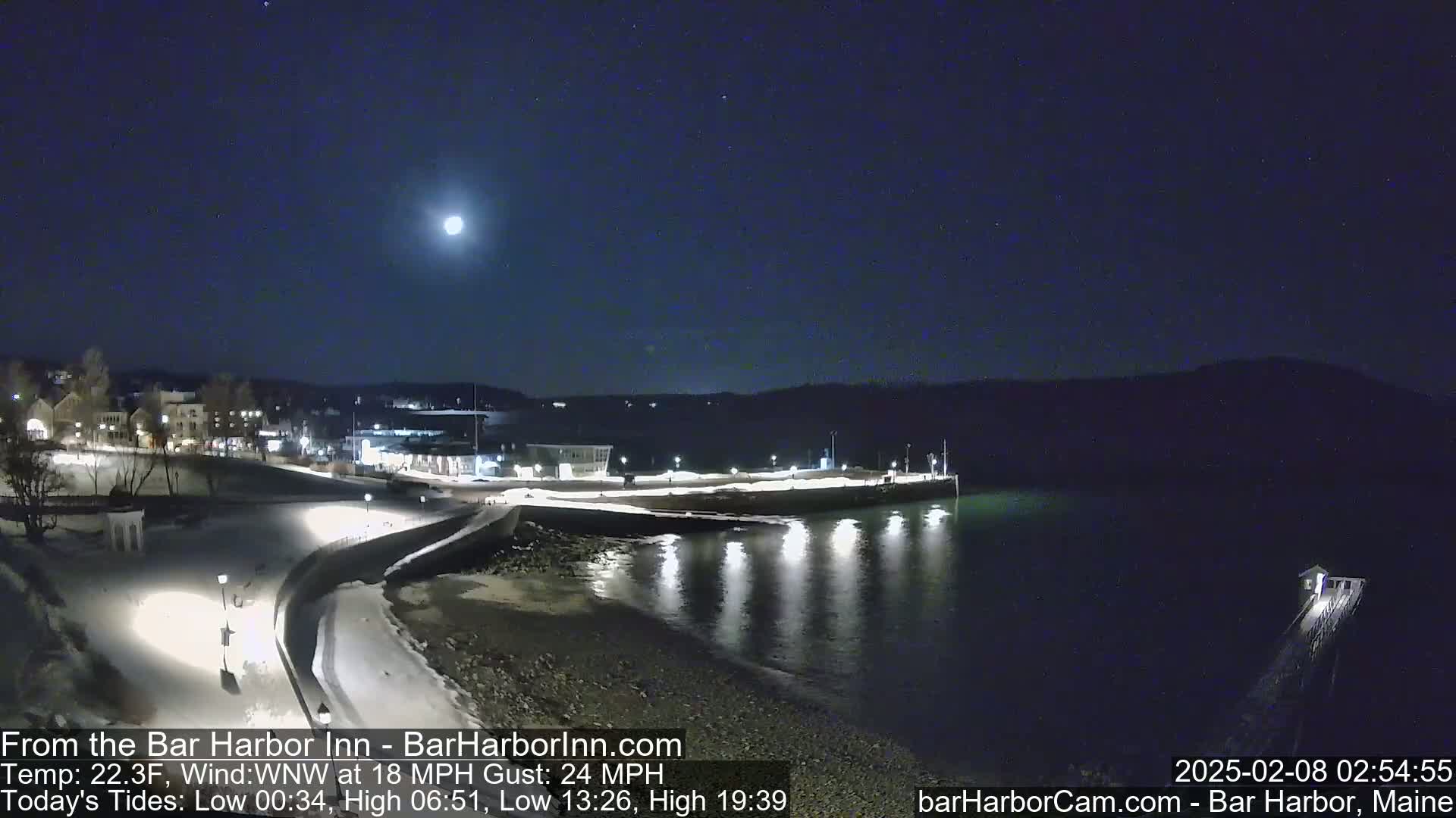 A moonlit nighttime scene shows a snow-covered waterfront with buildings in the distance under a clear sky.