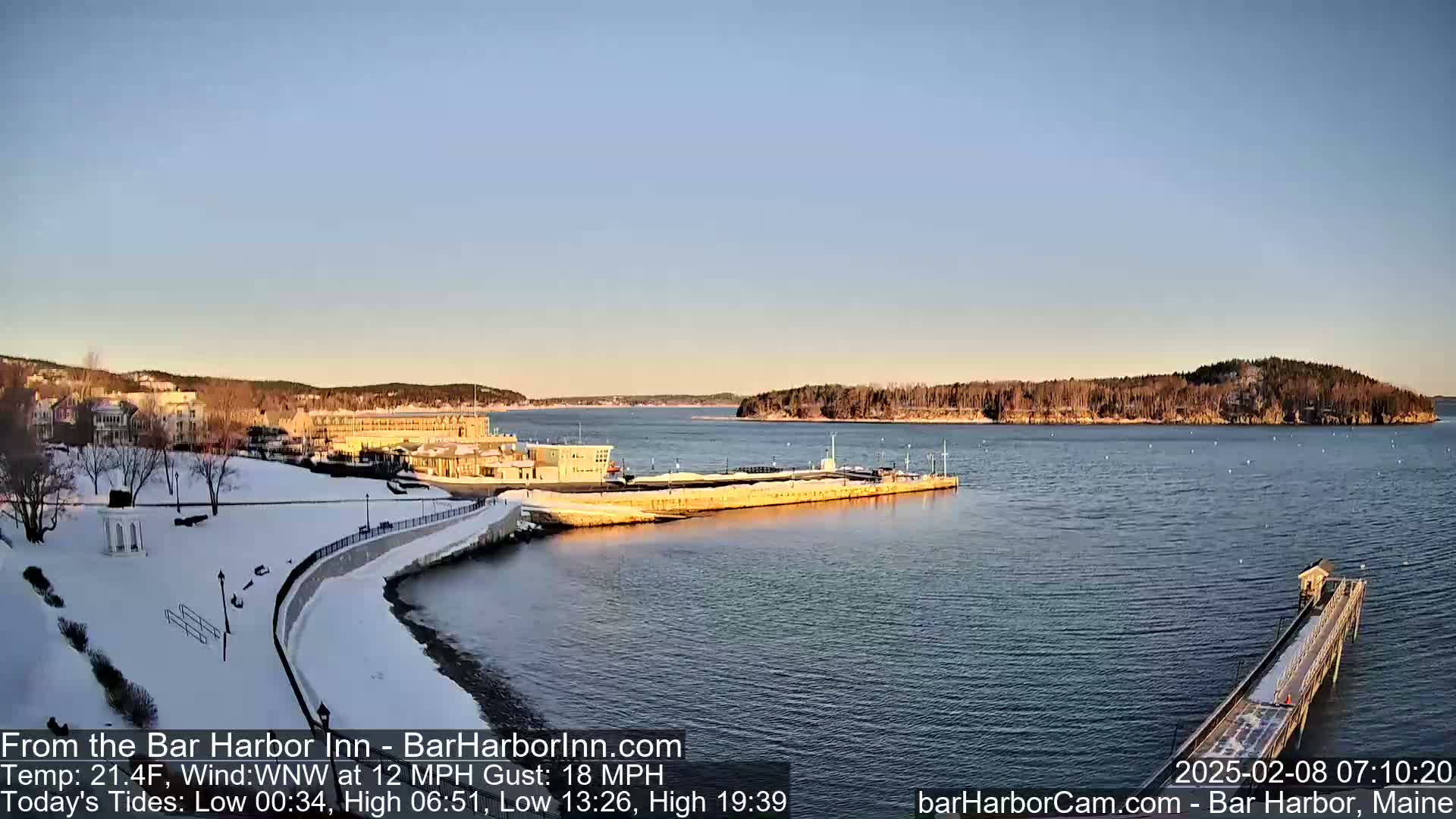 A snow-covered waterfront scene shows buildings, a partially frozen harbor, and a long pier extending into the calm water under a clear, bright sky.