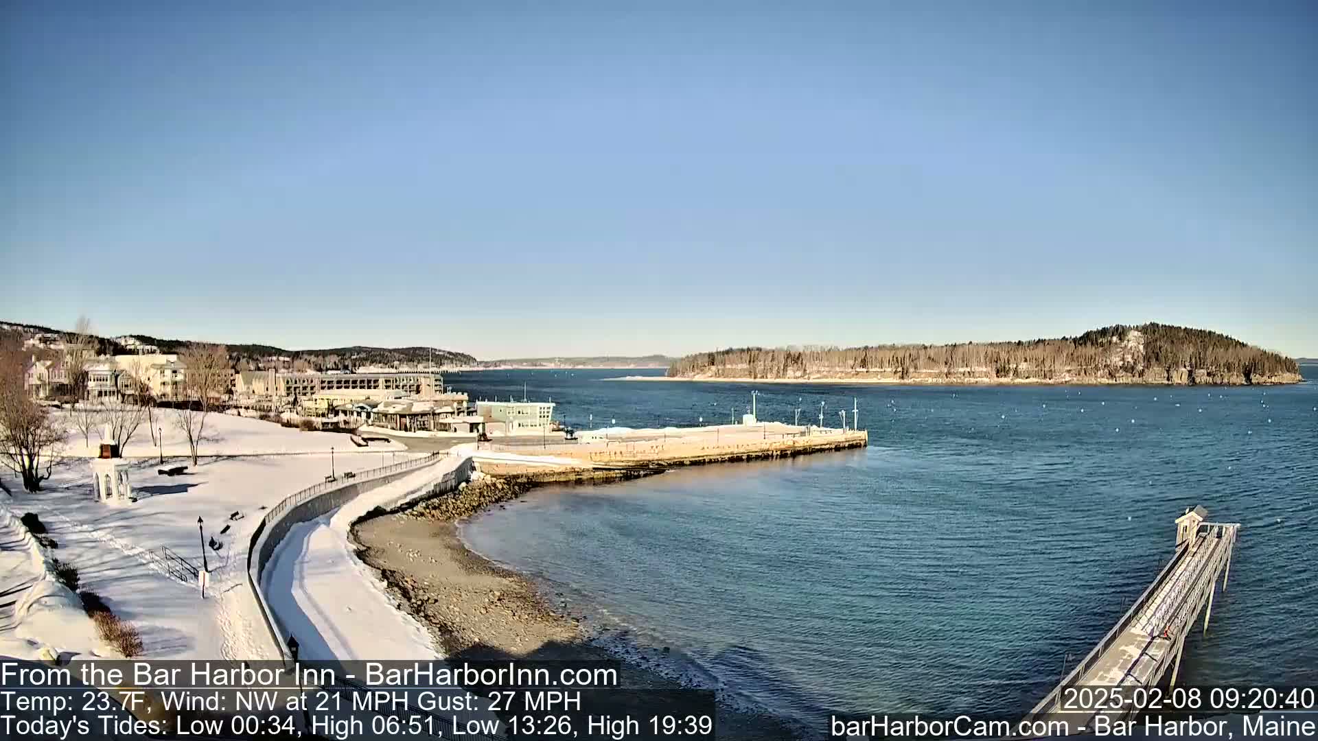 A snow-covered waterfront scene shows buildings, a partially frozen harbor, and a long pier extending into calm blue water under a clear, sunny sky.