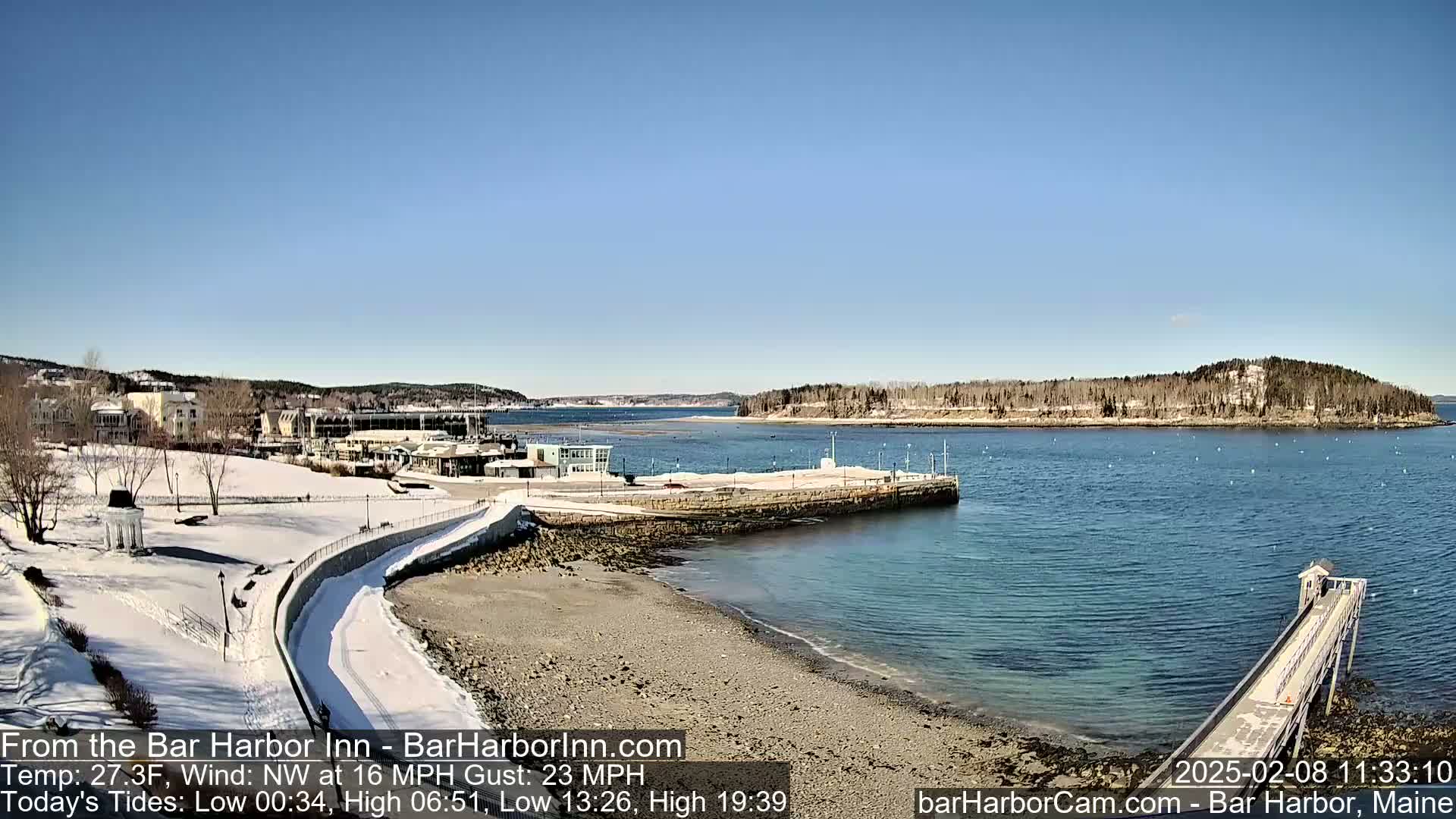 A snow-covered waterfront scene shows a partly frozen harbor with a long pier extending into the water under a clear blue sky.