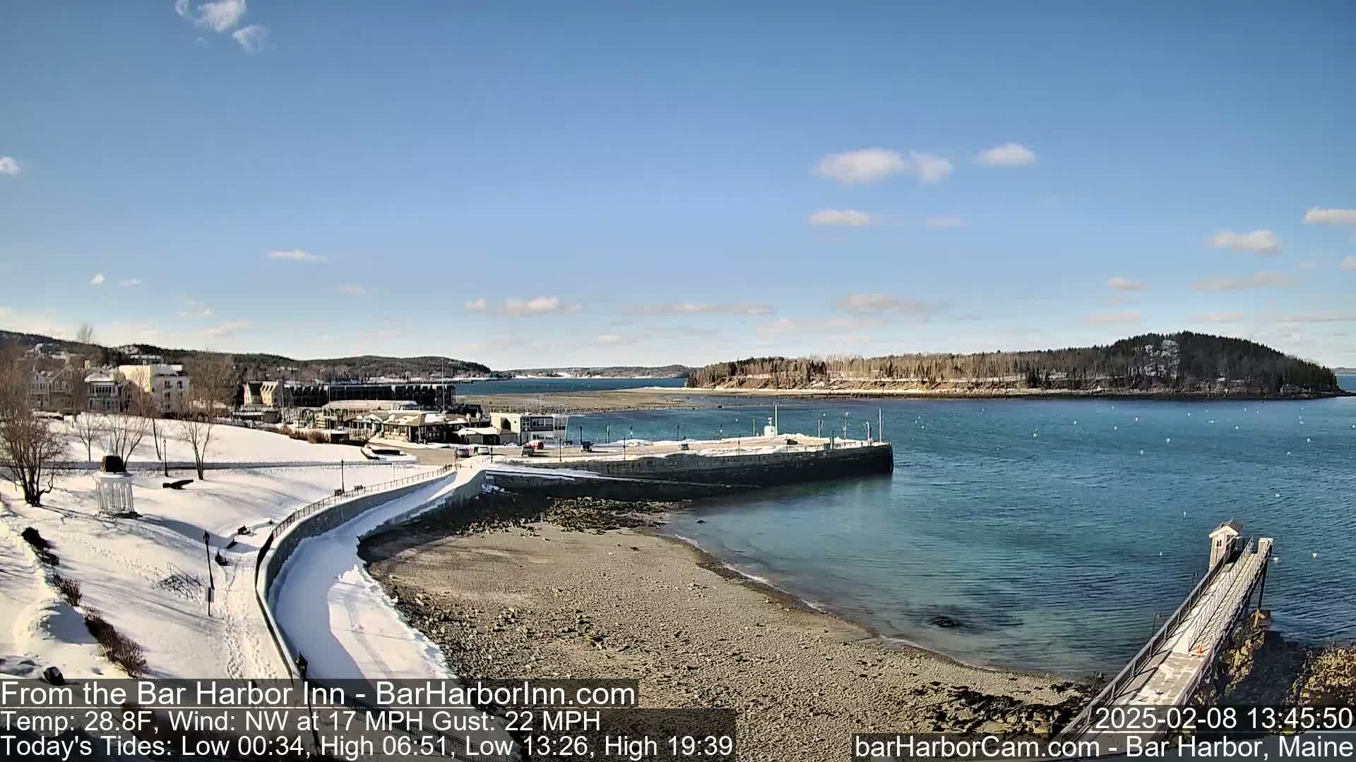 A snow-covered waterfront scene shows a partially frozen harbor with a small pier extending into the calm water under a mostly sunny sky.